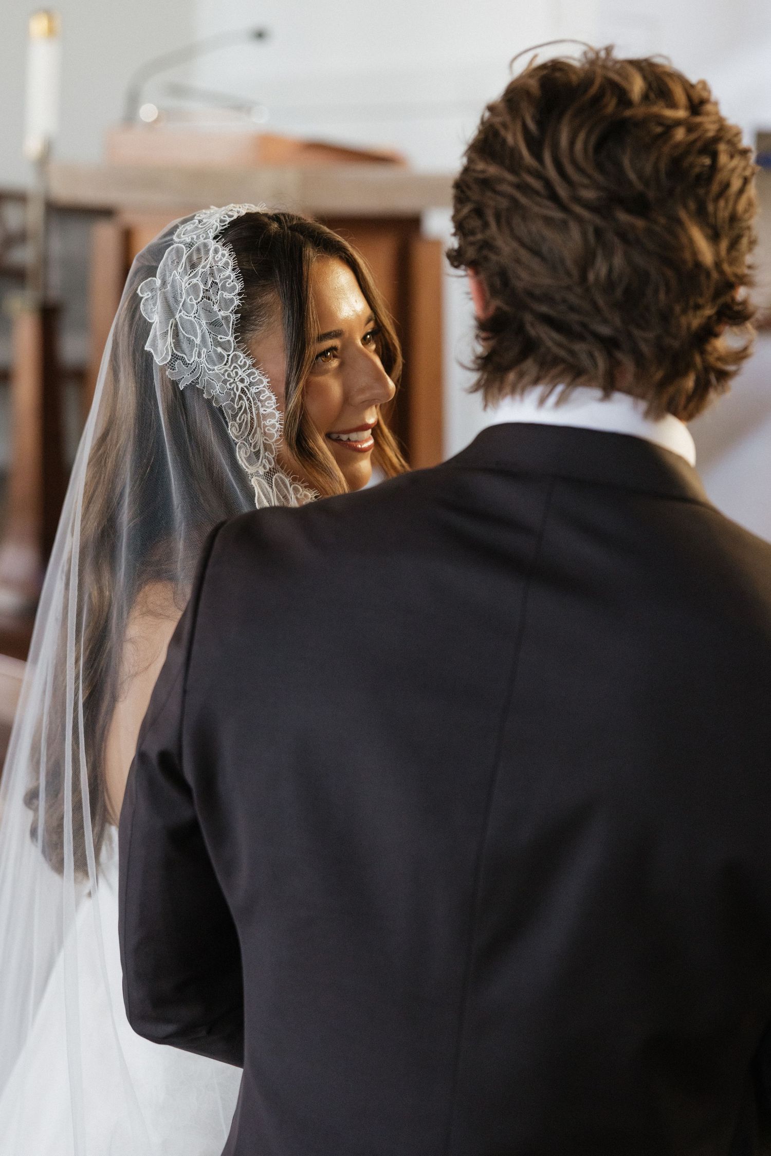 Smiling bride in lace veil gazes lovingly at groom during intimate church wedding ceremony.
