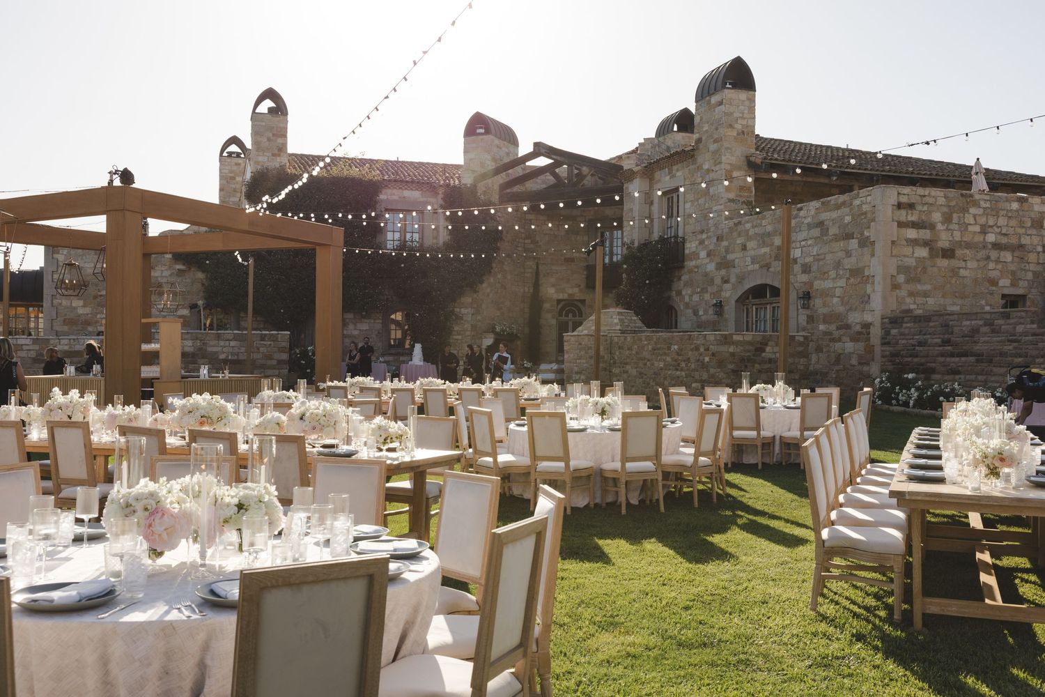 Outdoor wedding reception setup with white chairs and tables on grass beside historic stone building with string lights overhead.