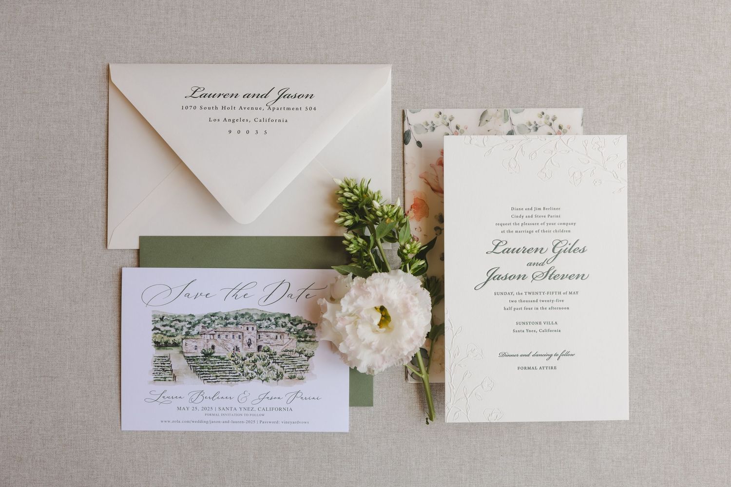 Elegant wedding invitation suite with white flowers and greenery on a neutral gray background.