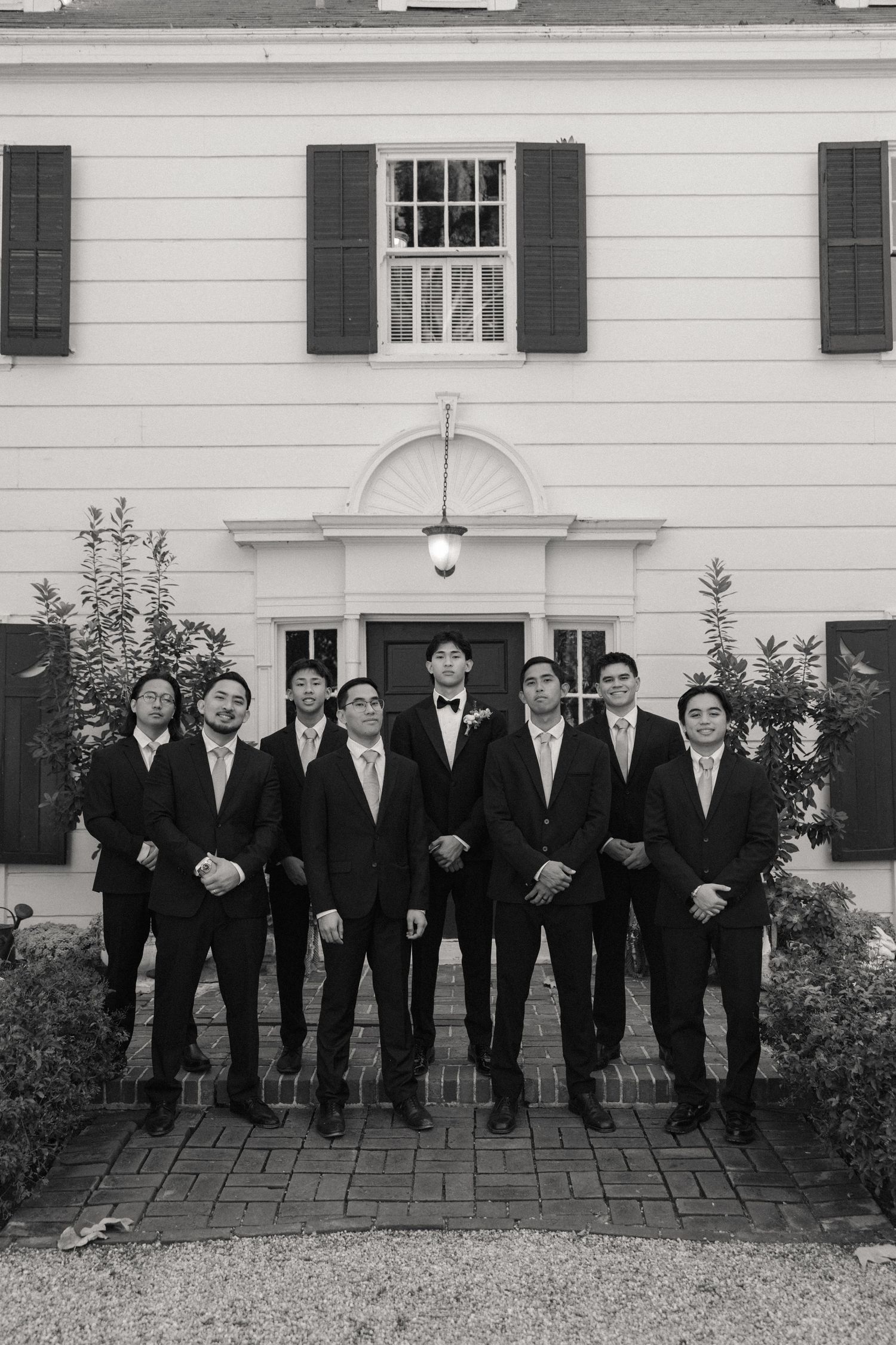 A line of groomsmen in black suits and ties stand in front of a white building with dark shutters.