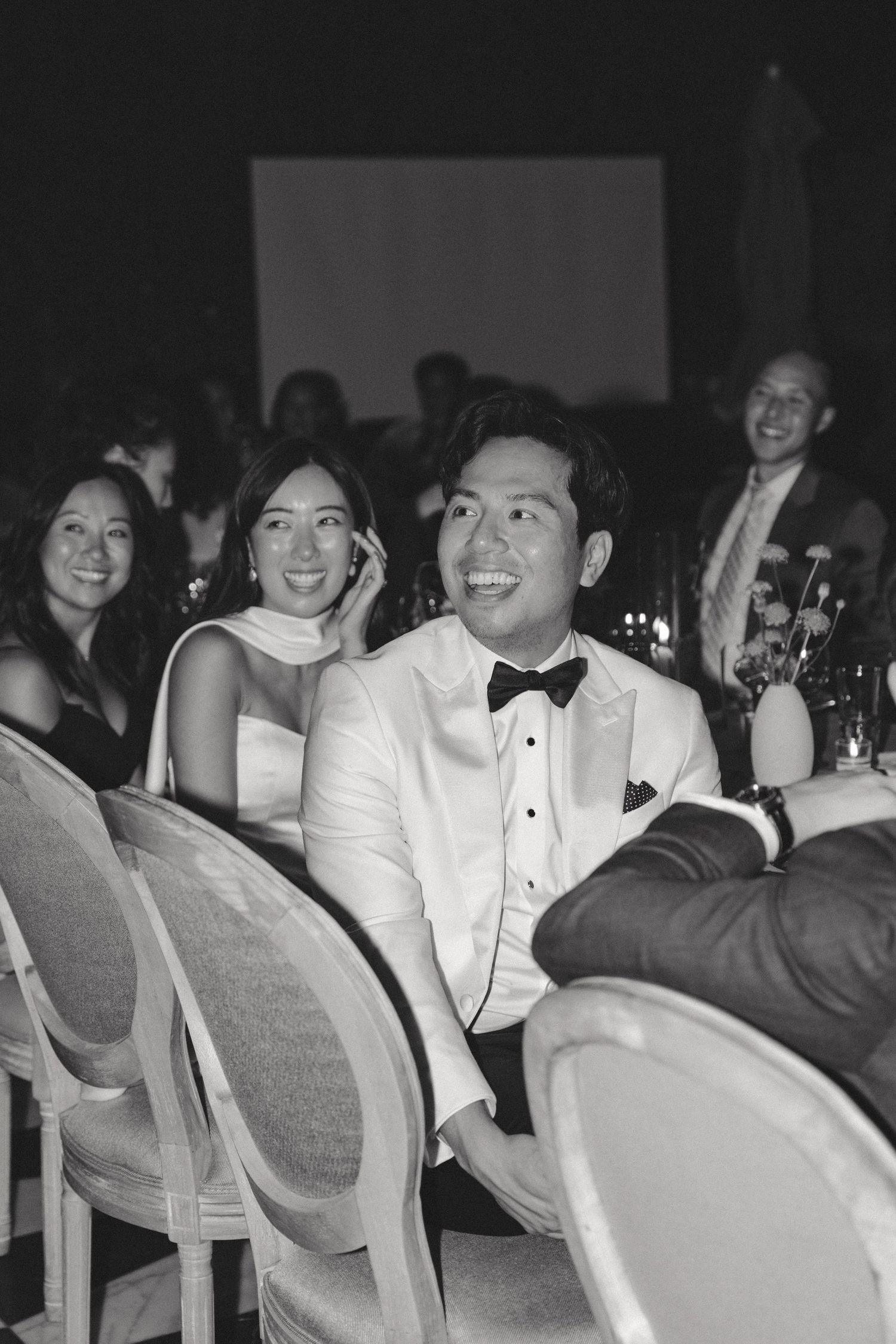 Wedding reception guests in formal attire seated in rows of chairs at an indoor venue.