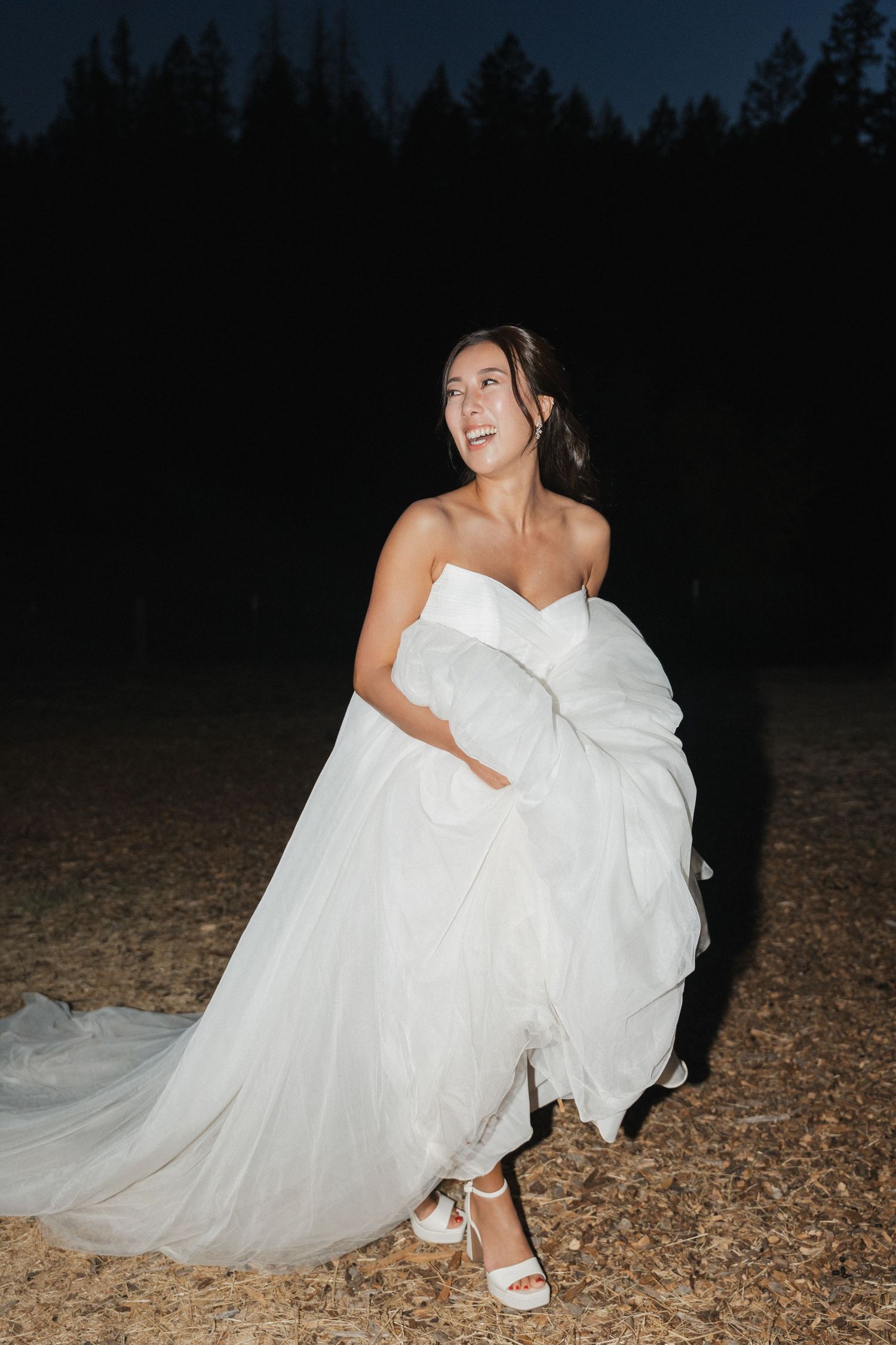 A bride in a strapless white gown laughs while holding her dress outdoors at night.