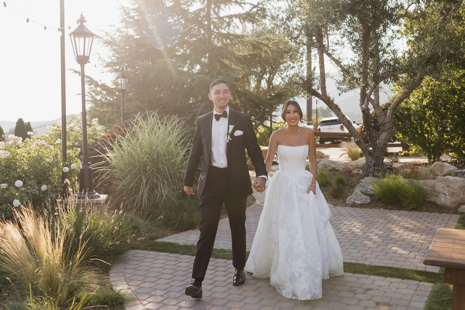 Newlyweds walking hand in hand through garden pathway at sunset.