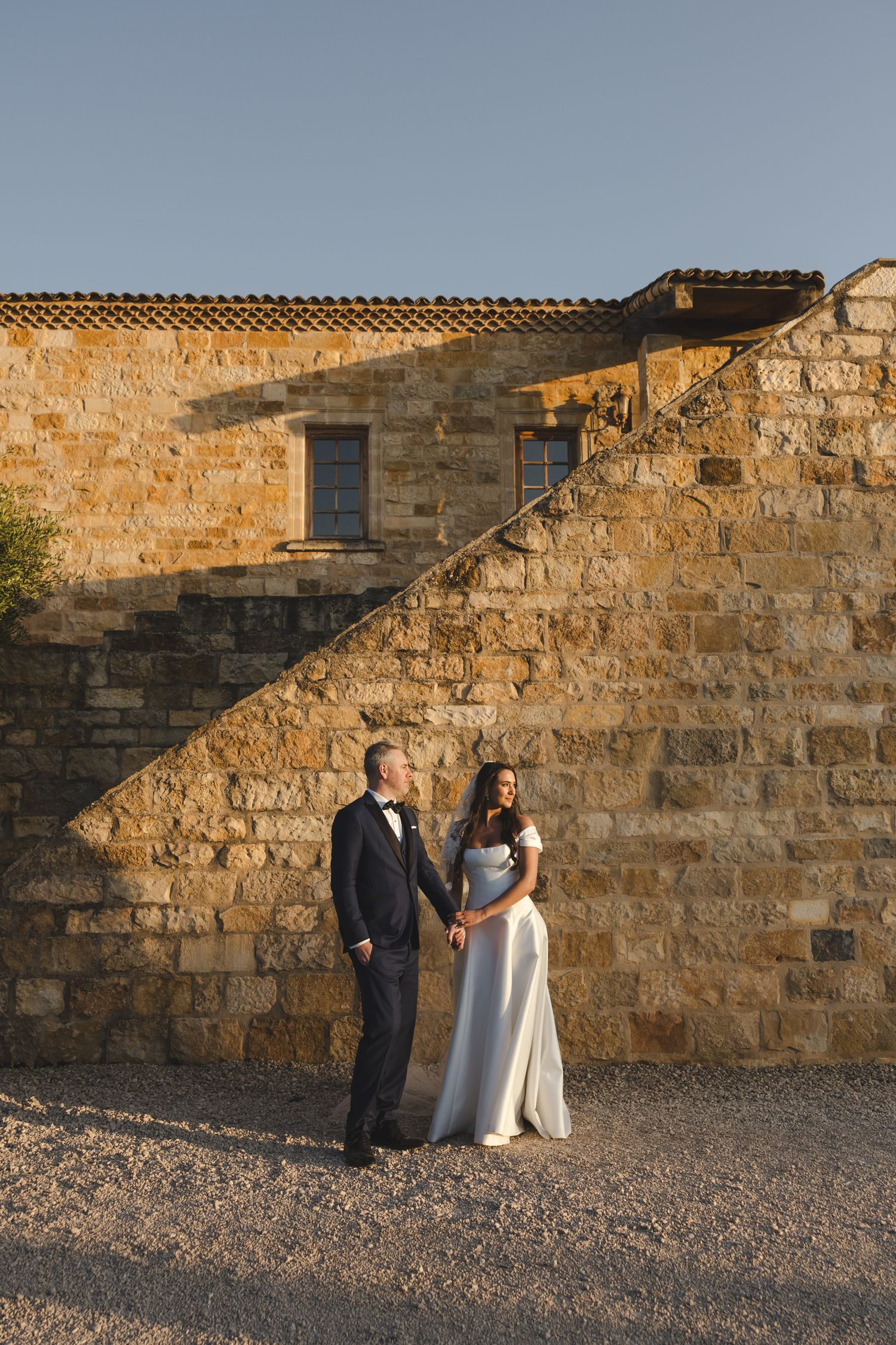 Elegant couple poses against rustic stone wall in warm evening light.