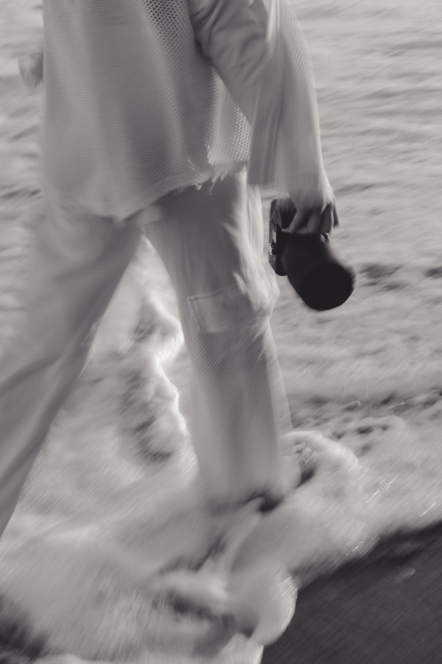 A black and white photo showing legs in white clothing and dark shoes walking on a textured surface.