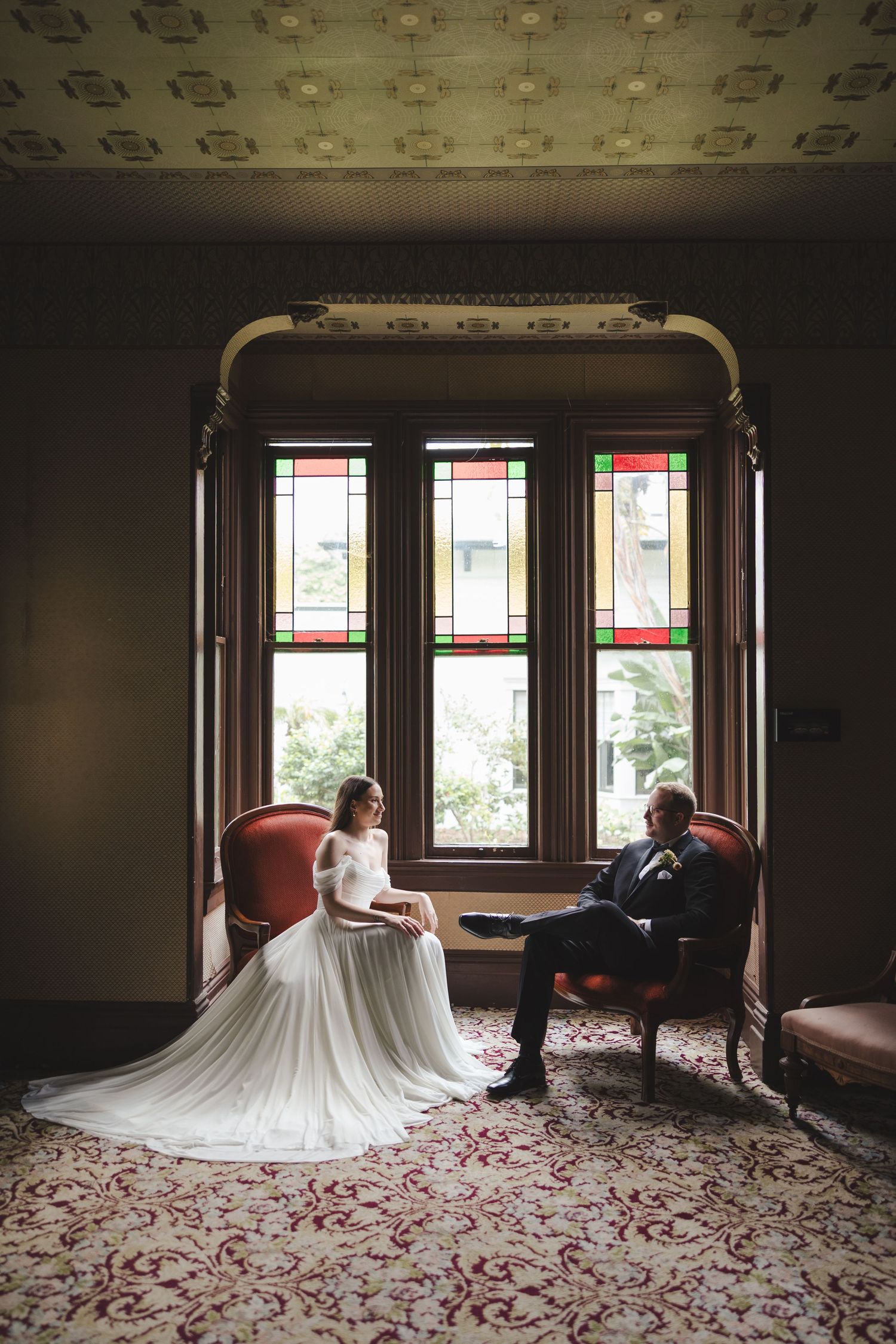Elegant couple seated by ornate stained glass windows in a vintage room with decorative ceiling and patterned carpet.
