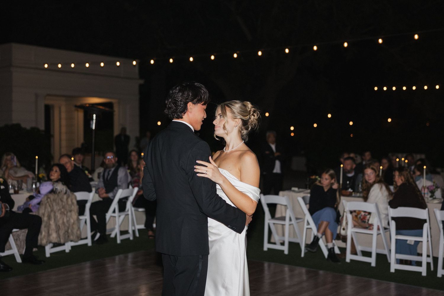 Evening outdoor wedding reception setup with string lights and white chairs under dark sky.