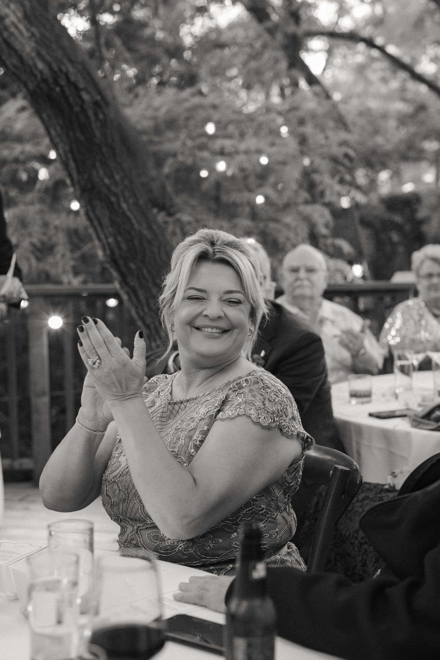 Black and white photo of a smiling blonde woman in lace dress clapping at an outdoor wedding reception.
