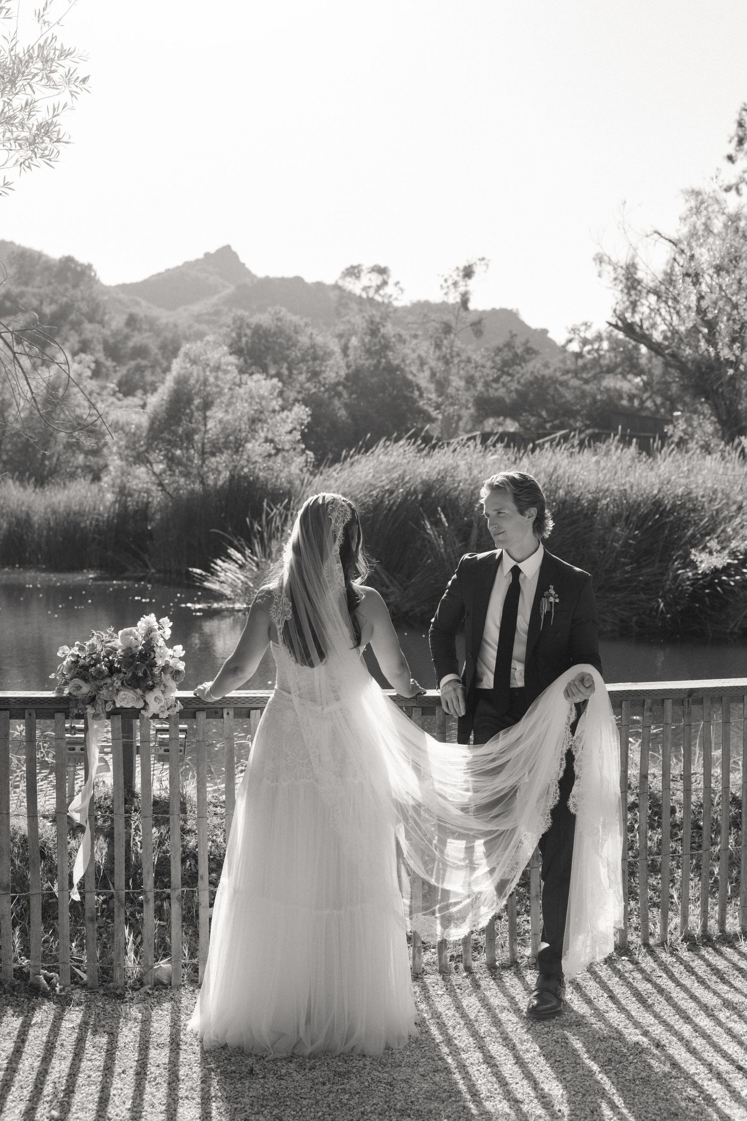 Black and white photo of bride and groom holding hands on wooden bridge with mountain lake backdrop.