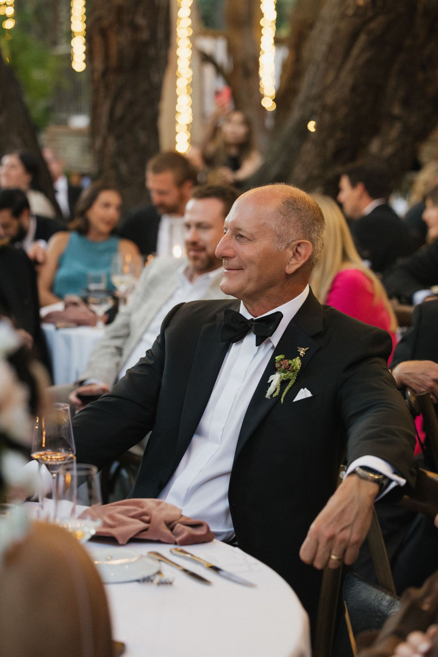 Elegant man in tuxedo with boutonniere smiles at outdoor wedding reception under twinkling lights.