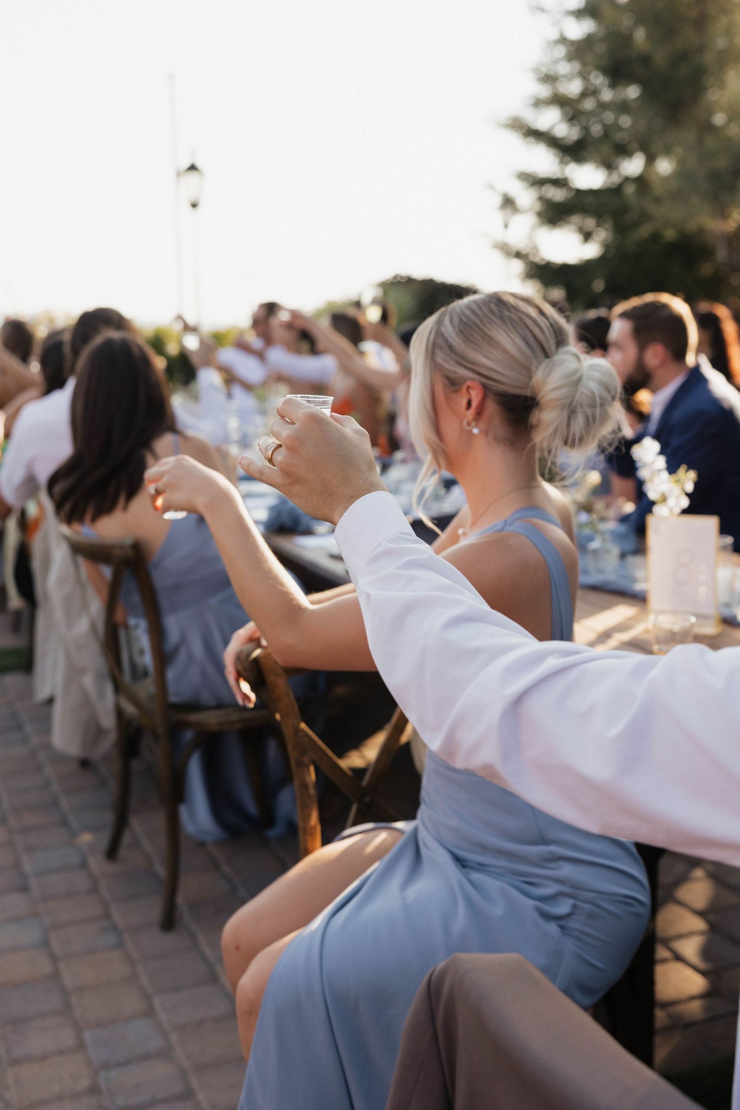Wedding guests seated at outdoor tables applaud during golden hour celebration.