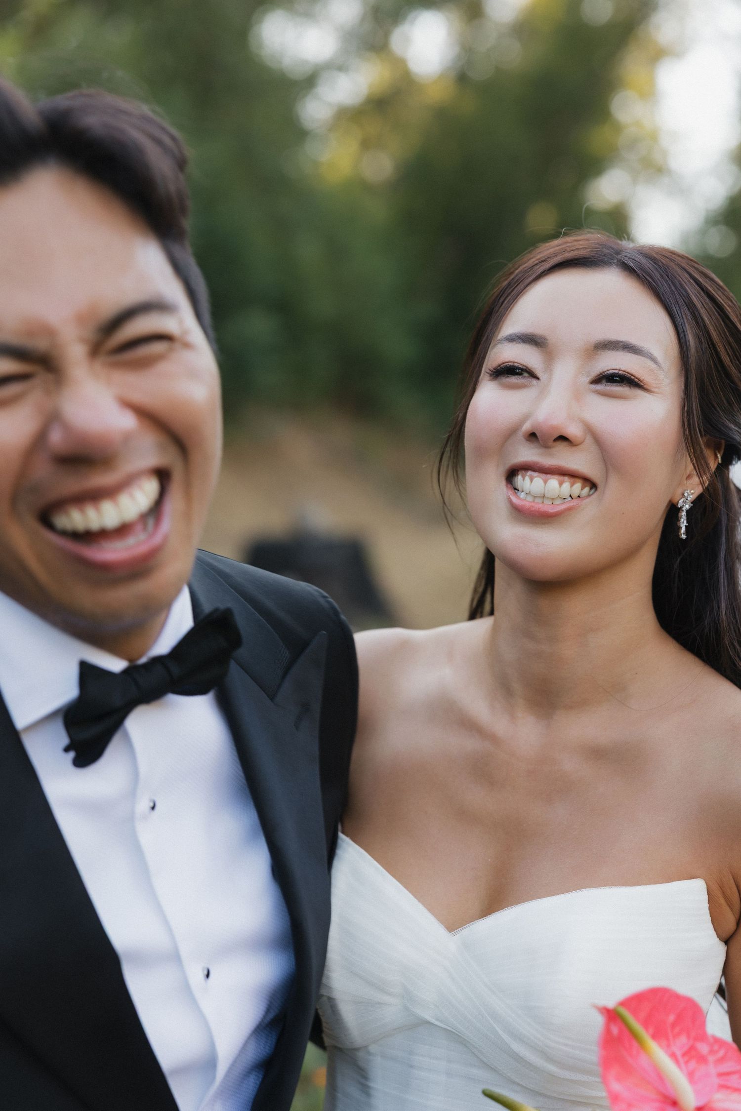 A wedding couple shares joyful laughter while holding a pink bouquet outdoors.