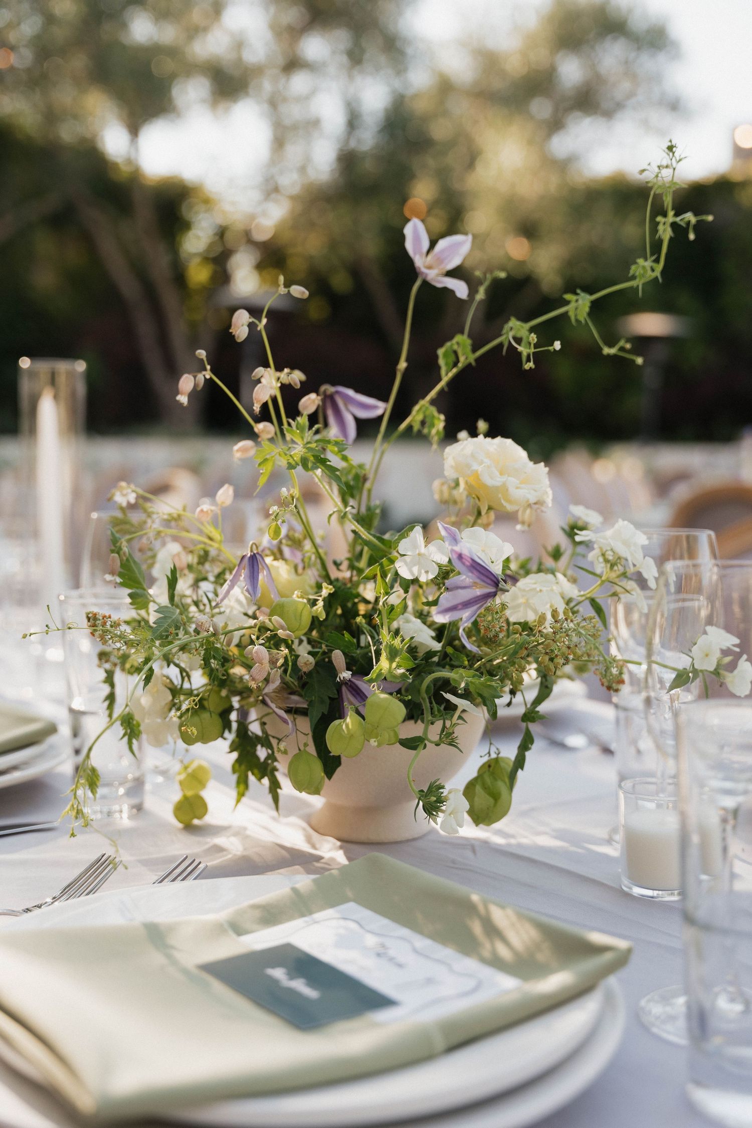 Elegant white floral centerpieces and place settings adorn tables at an outdoor wedding reception.