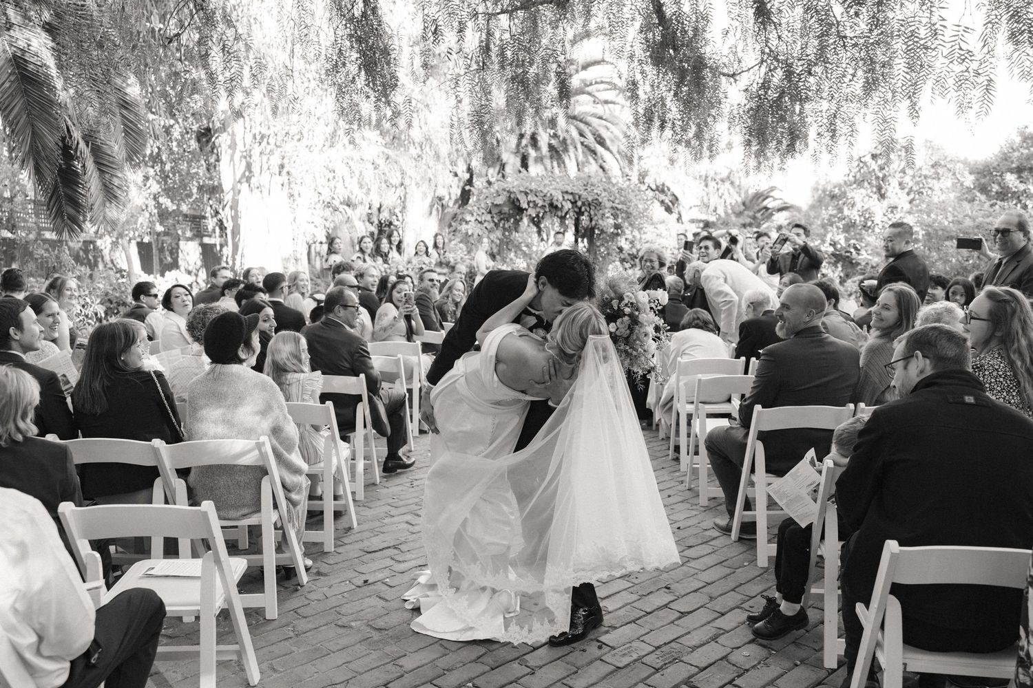 A romantic black and white photo of a couple sharing their first dance at an outdoor wedding ceremony with guests seated.