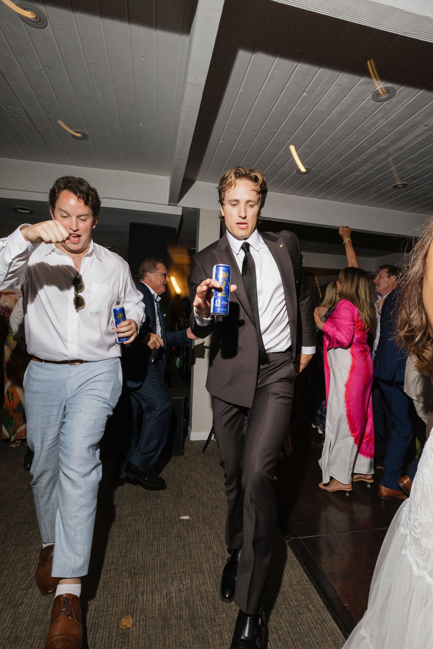 Groom in dark suit dancing with beer can at lively wedding reception.