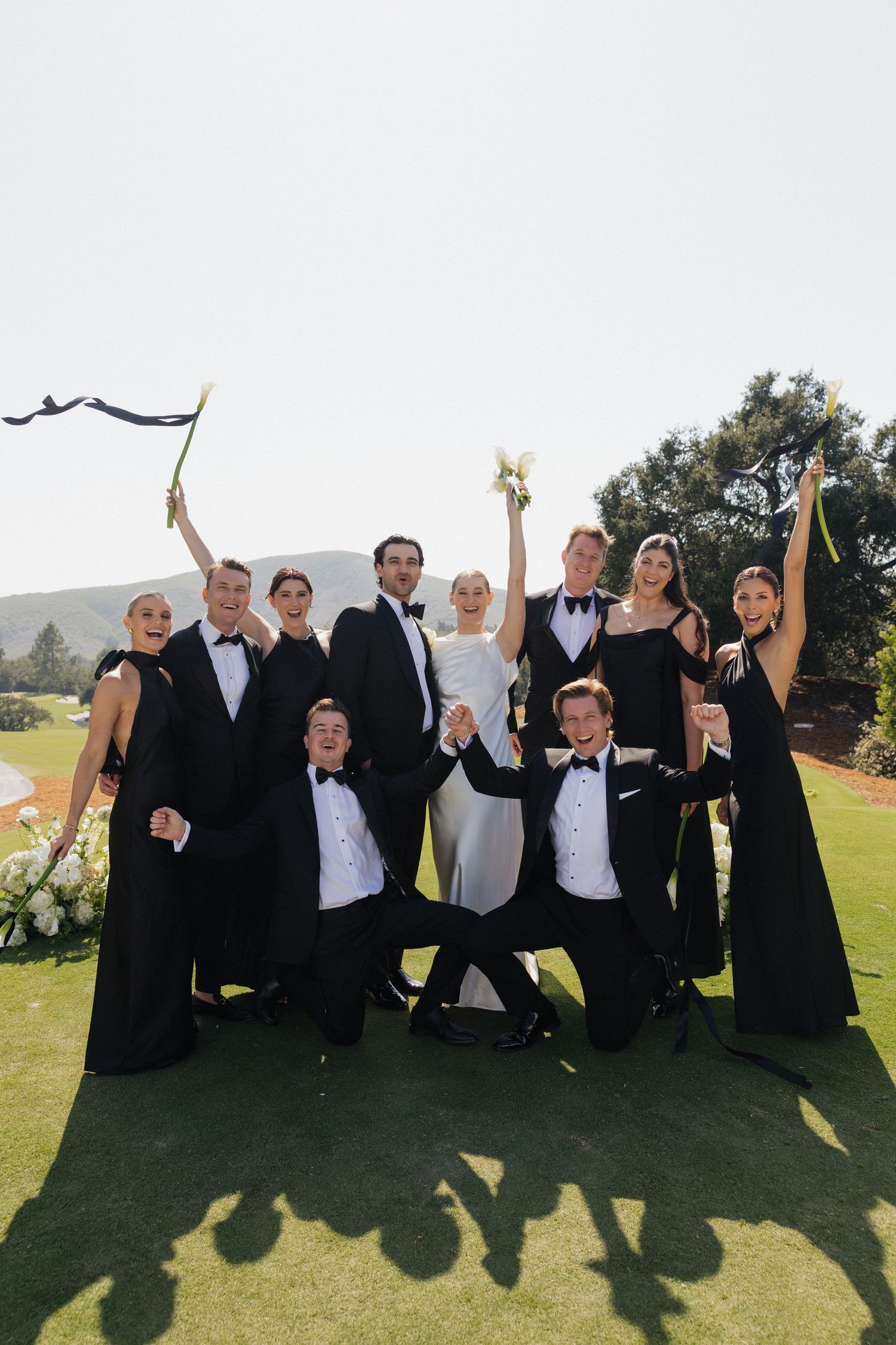 Wedding party in black formal wear celebrates outdoors on green lawn, bride in white gown at center, all cheering joyfully.