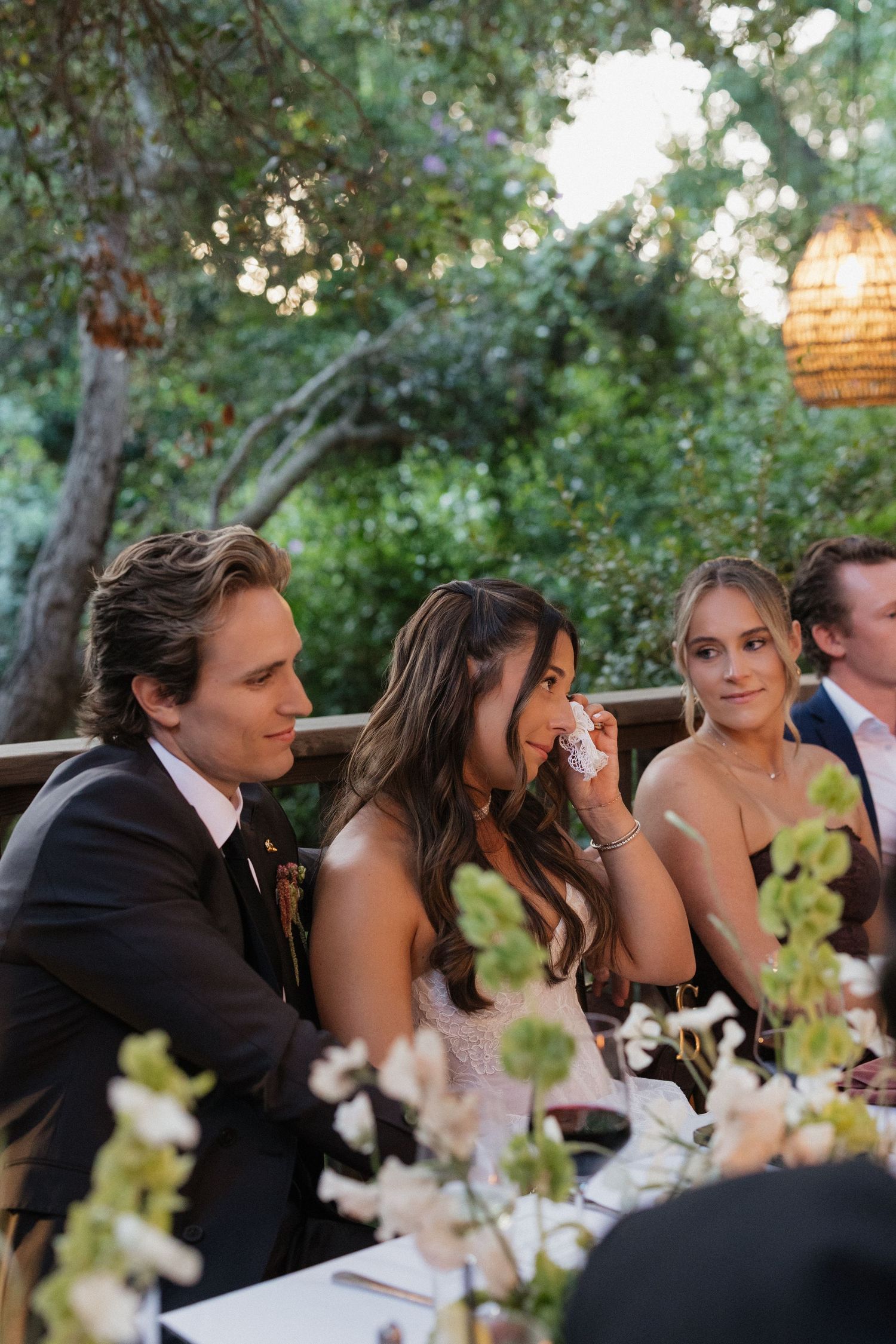 Bride wiping tears at outdoor wedding reception dinner table surrounded by guests.