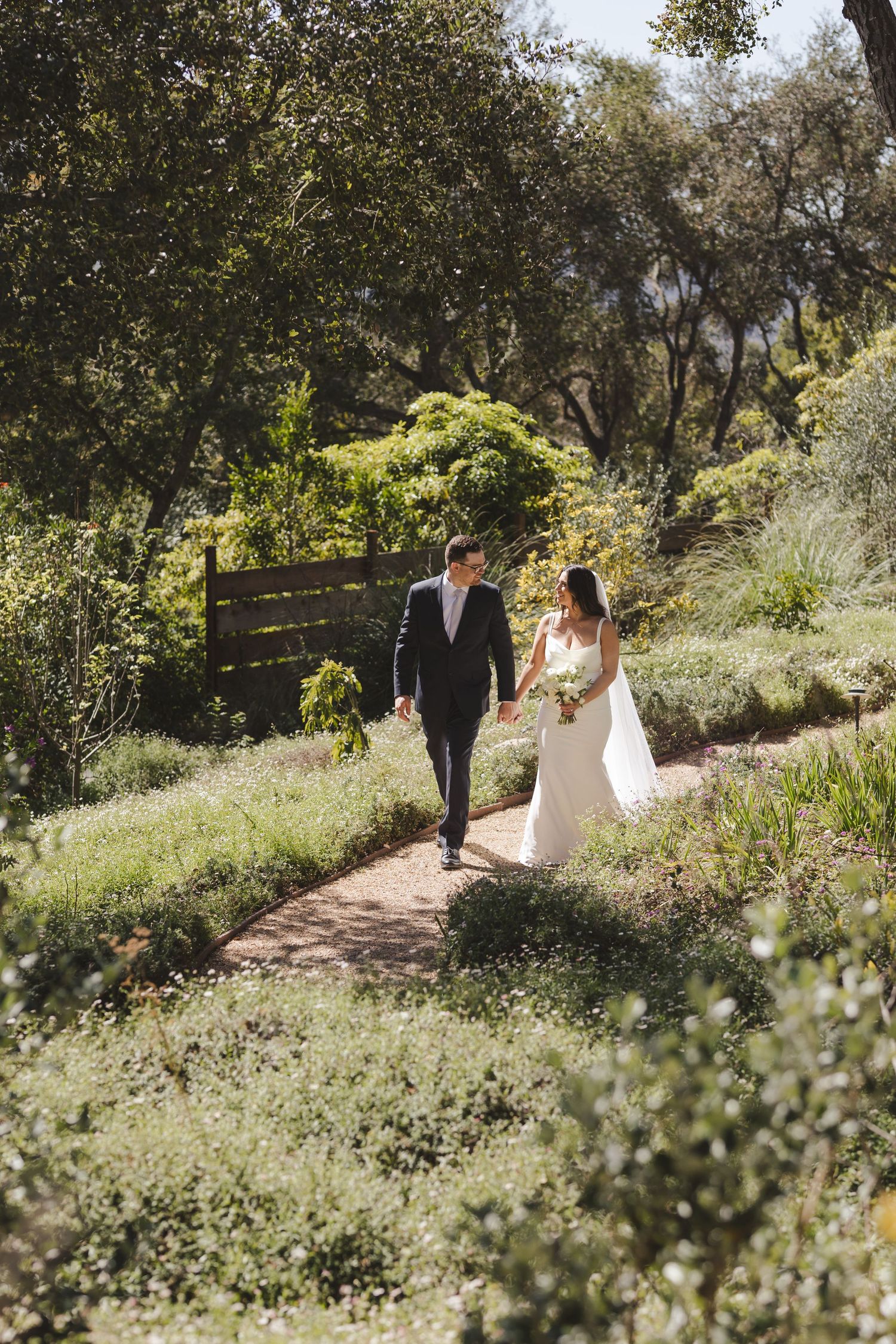 A couple walks down a winding garden path surrounded by lush greenery and trees on a sunny afternoon.