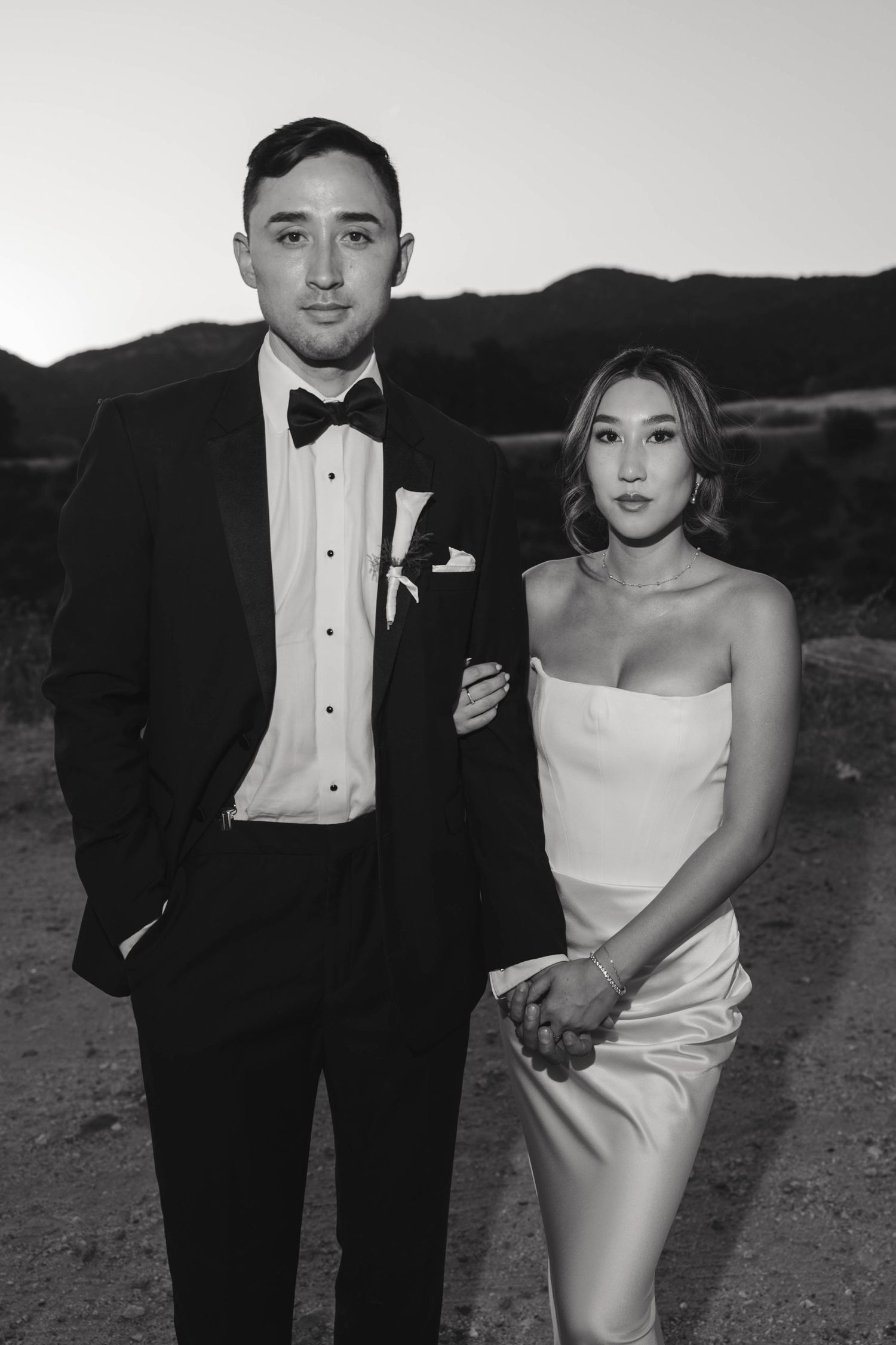 A formal black and white portrait of a couple in evening wear against a mountain landscape at dusk.