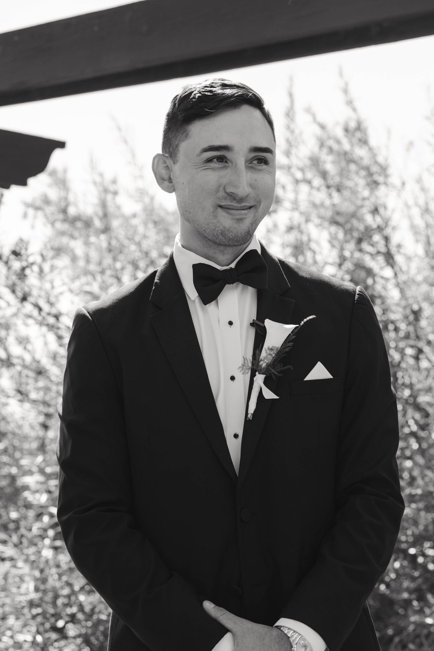 Groom in classic black tuxedo with white pocket square smiles during outdoor wedding photoshoot.