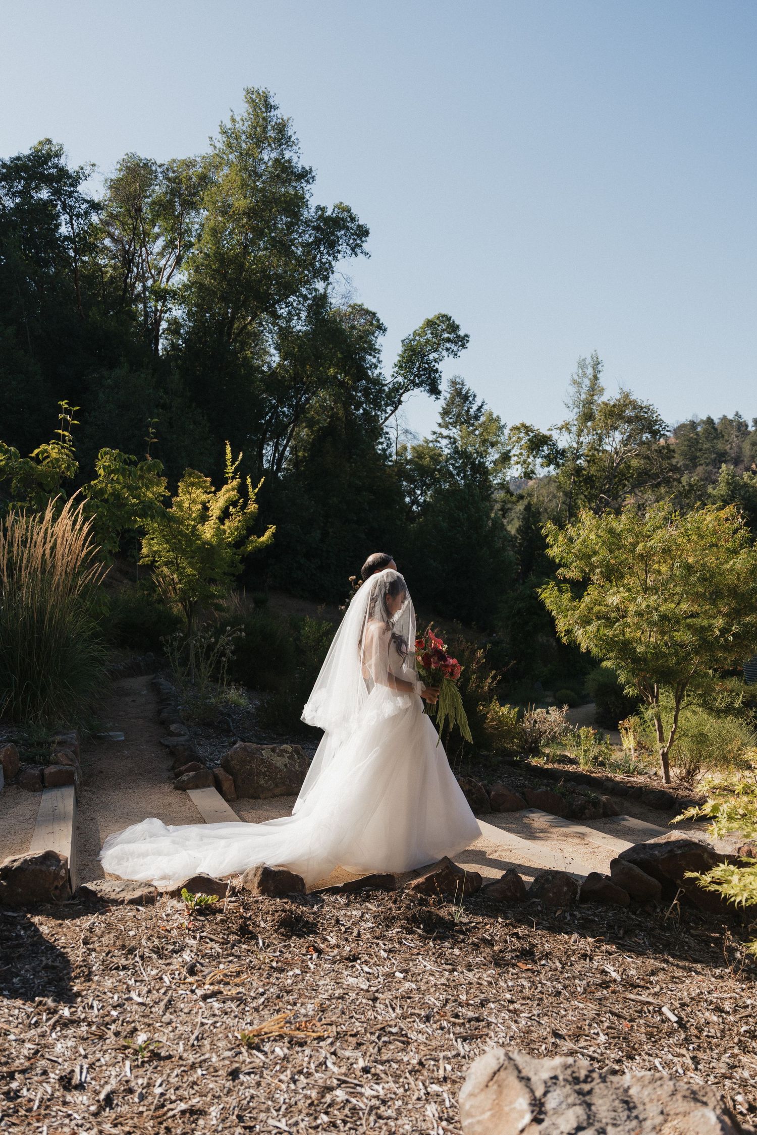 A bride in a flowing white wedding dress poses on a rocky hillside surrounded by trees and natural scenery.