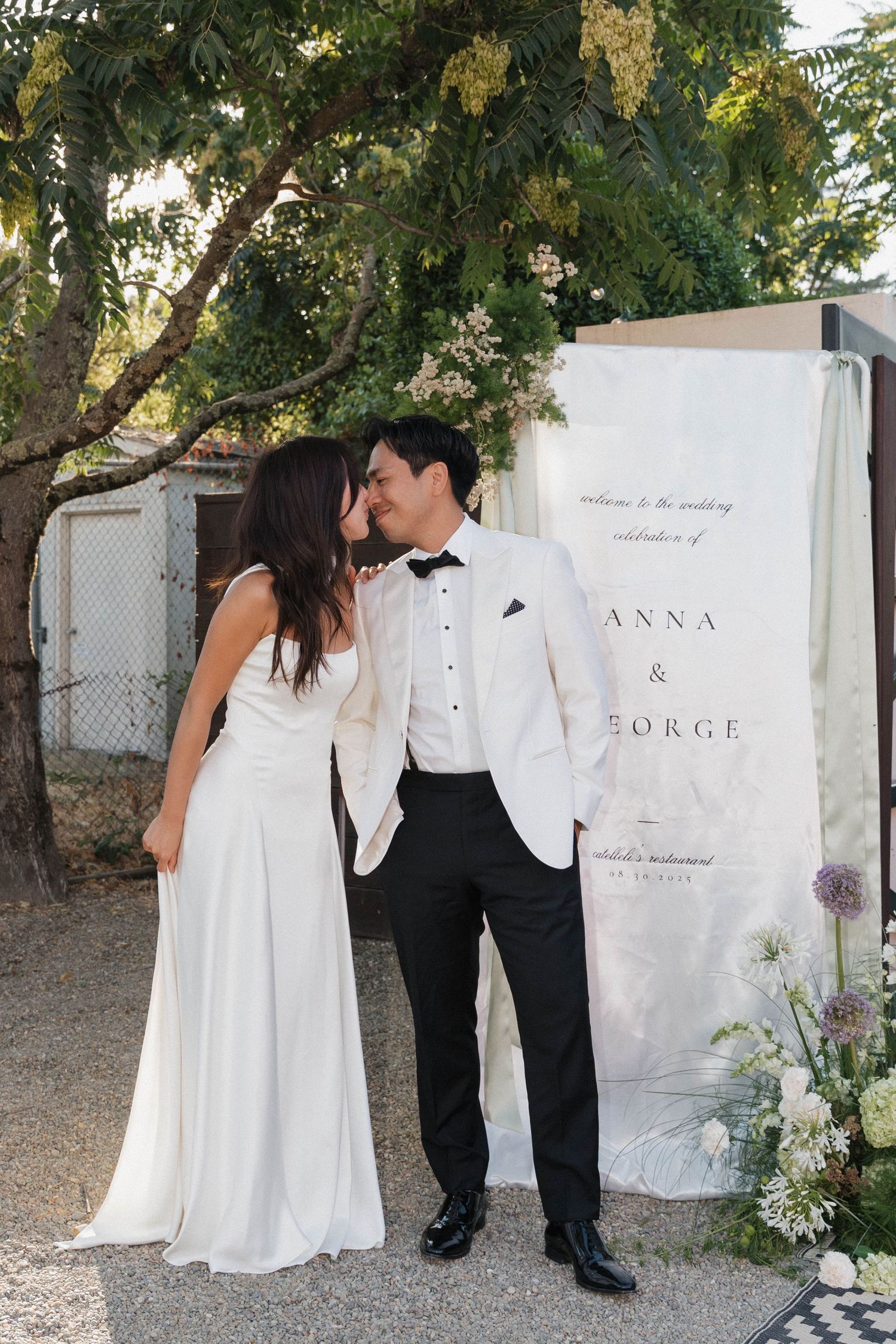 Wedding couple poses against a white backdrop with elegant floral decorations and signage.