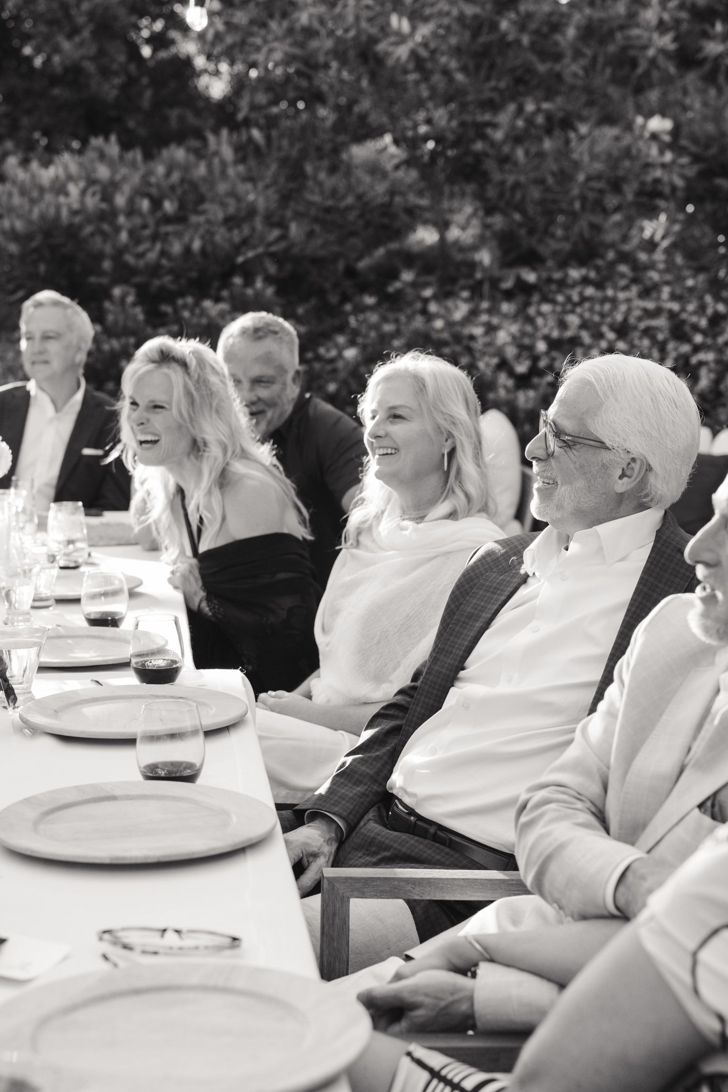 Black and white candid shot of people laughing at an outdoor dinner party with empty plates on a long table.