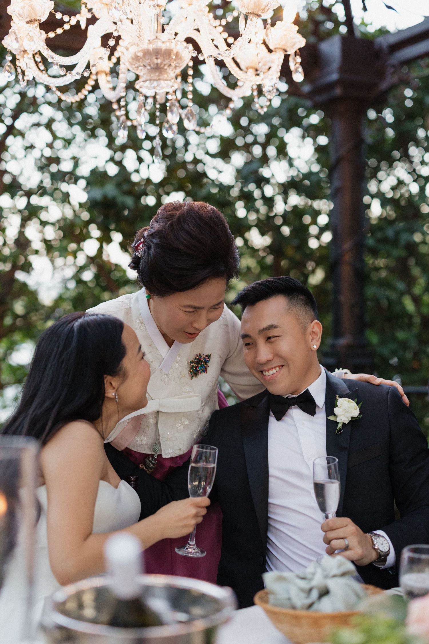 A joyful wedding reception moment with a bride, groom in black tuxedo, and older woman sharing champagne under crystal chandeliers.