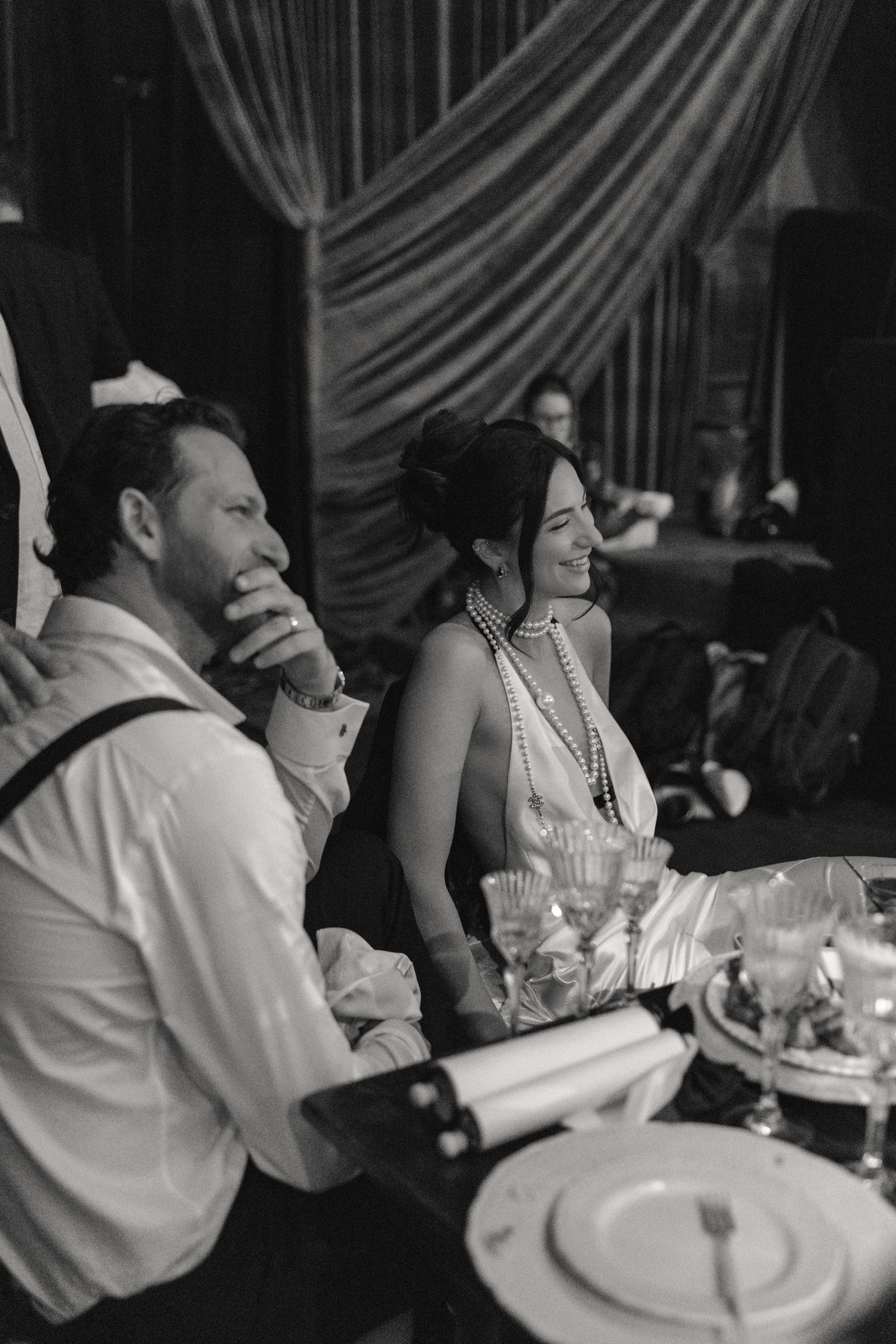 A couple sits at an elegant dinner table, with the woman wearing pearls and the man in a white dress shirt.