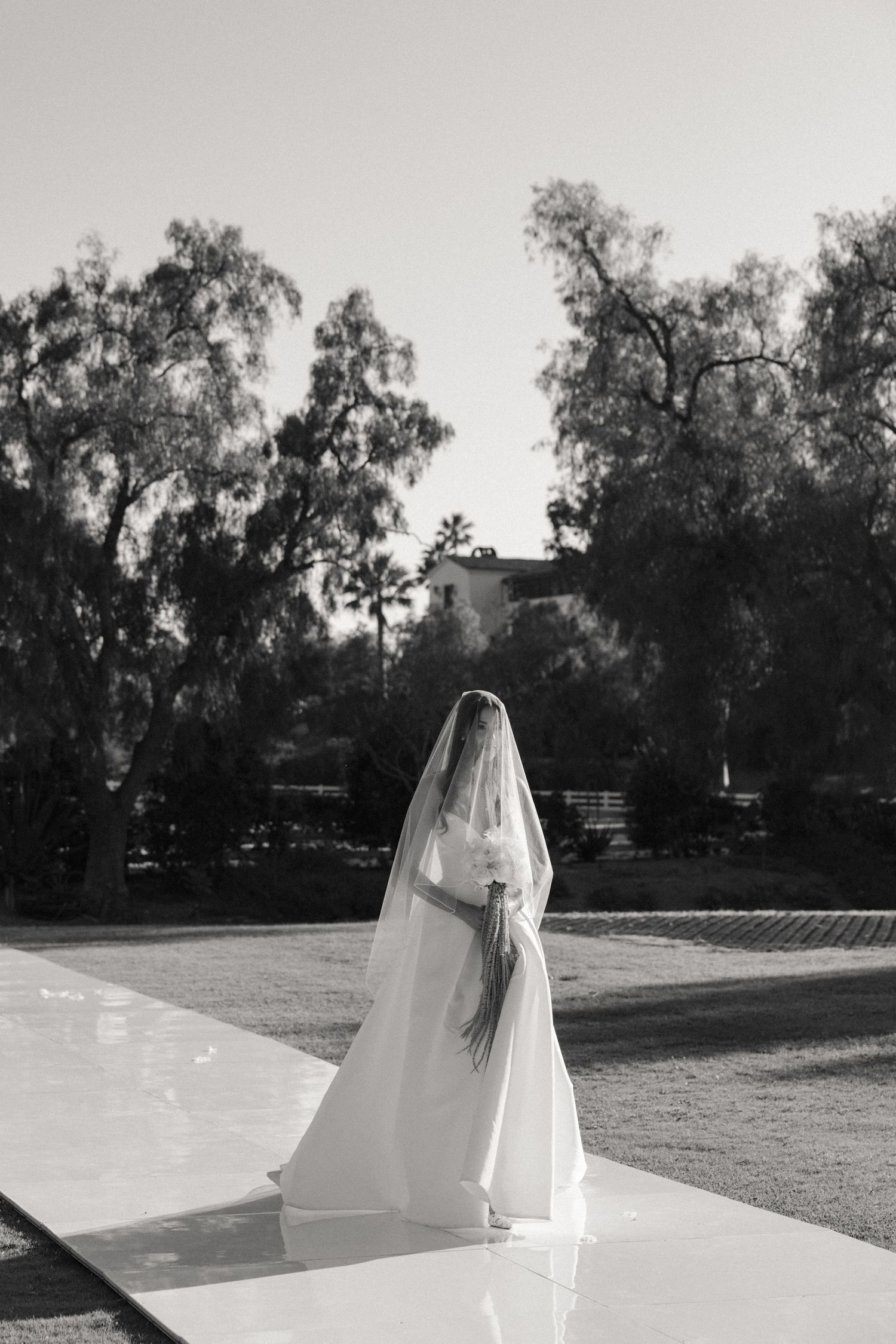 A figure in a flowing white wedding veil stands in a garden setting captured in black and white photography.