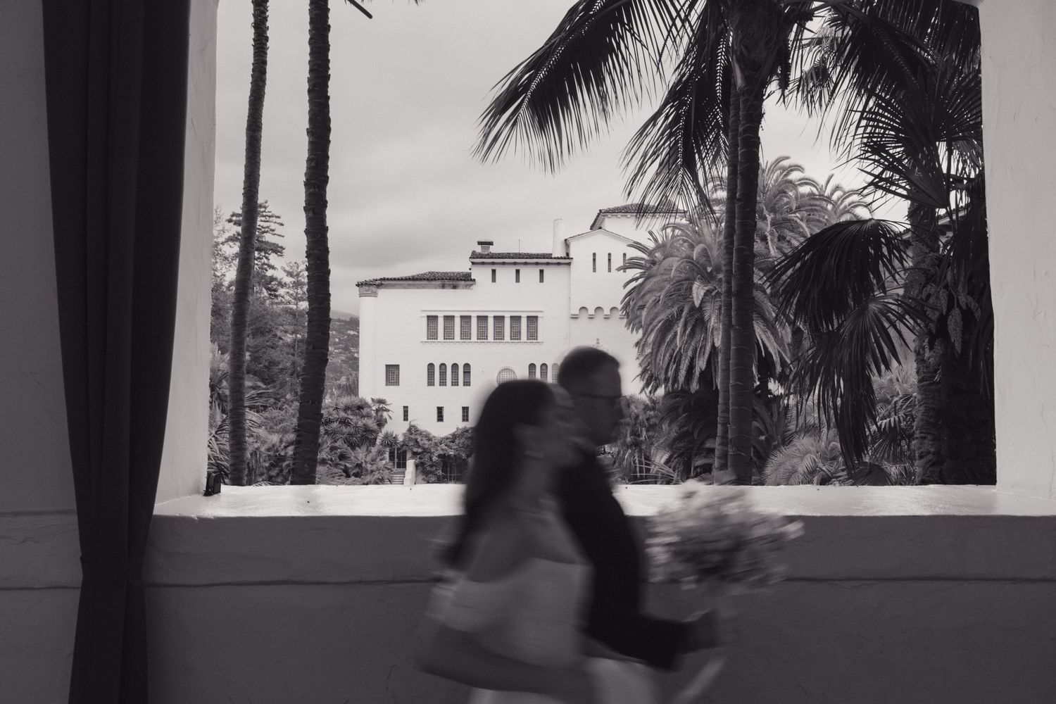 A blurred figure walks past a window overlooking a Mediterranean-style building with palm trees in black and white.