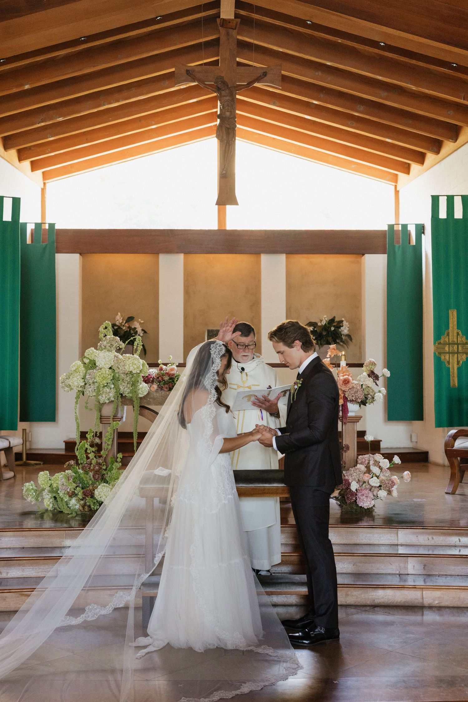 Bride and groom exchange vows at church altar with wooden ceiling, cross, and green banners during wedding ceremony.