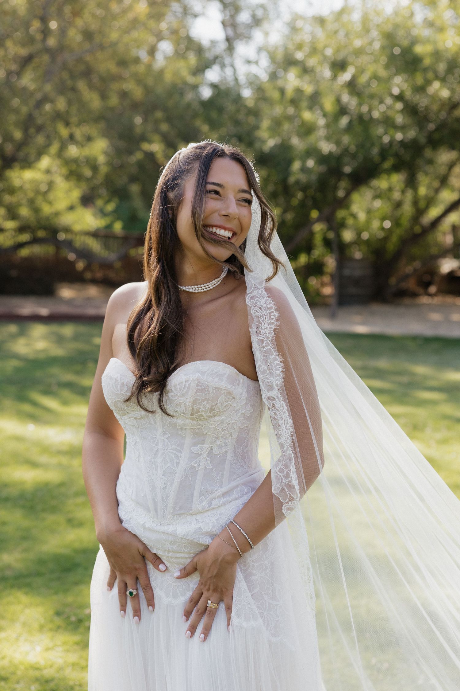 Radiant bride in strapless lace gown and flowing veil laughs joyfully outdoors on wedding day.