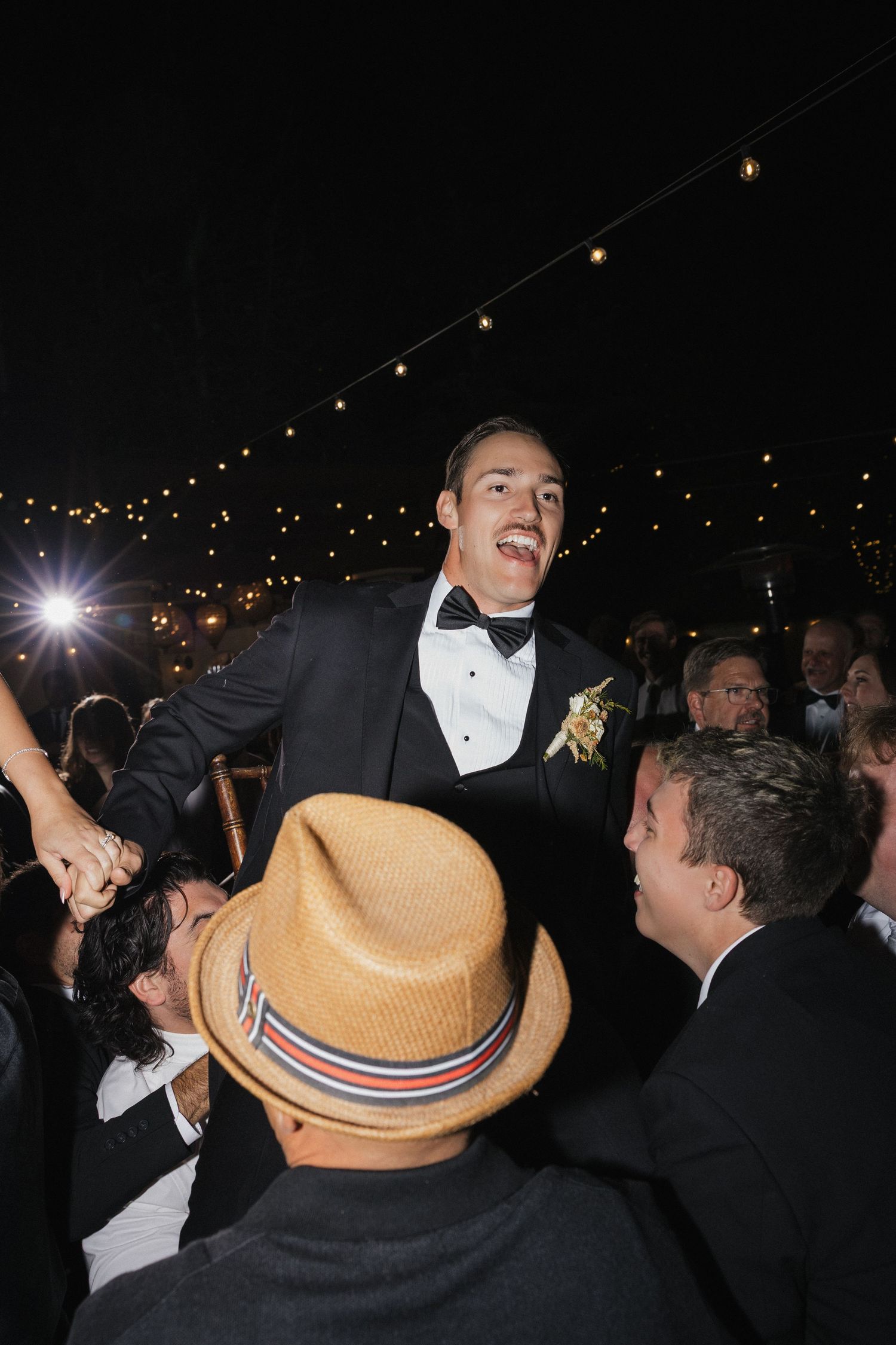 Wedding guests celebrate on a dance floor at night under string lights with someone wearing a straw hat.