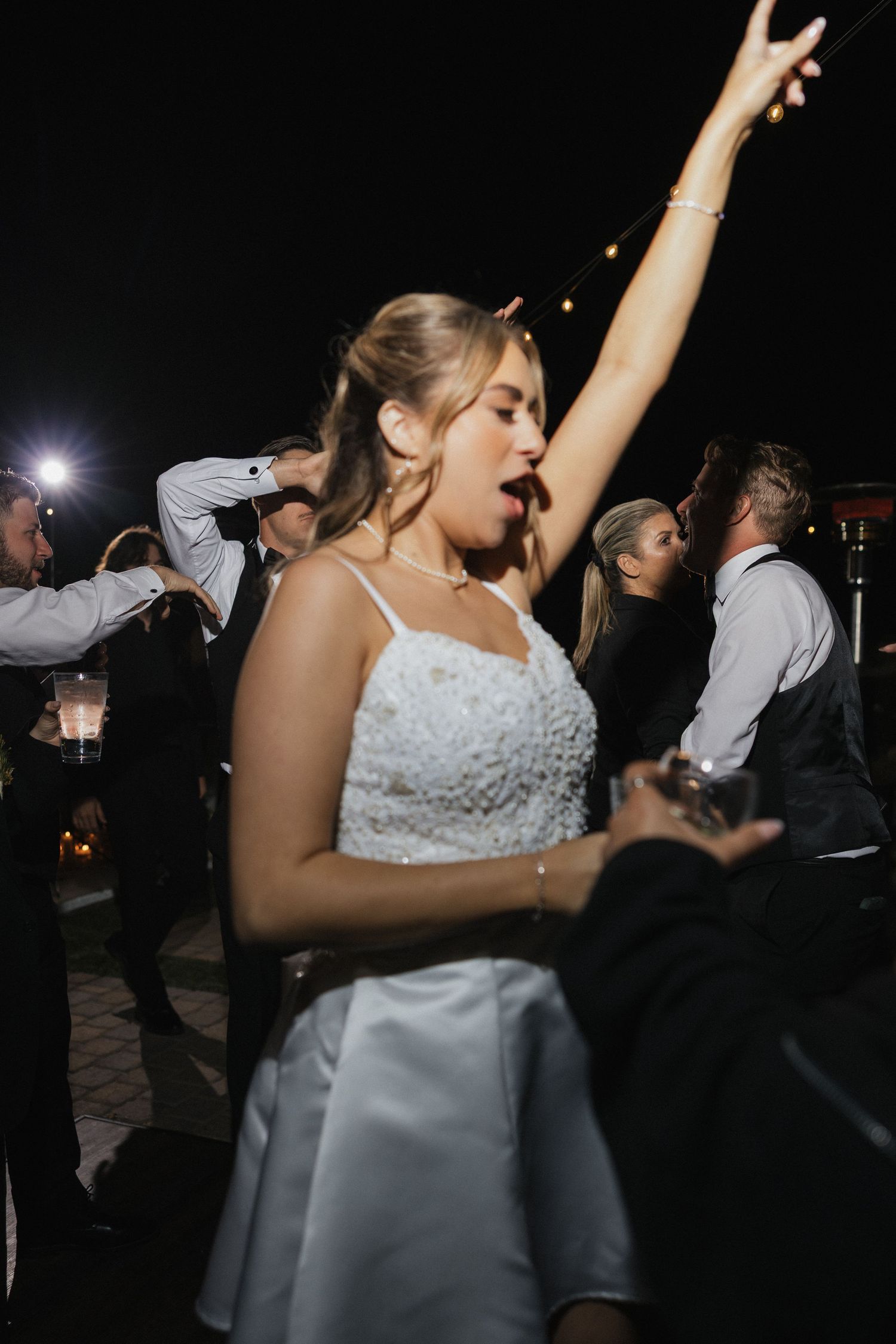 Wedding guests dance enthusiastically at an outdoor nighttime reception under string lights.