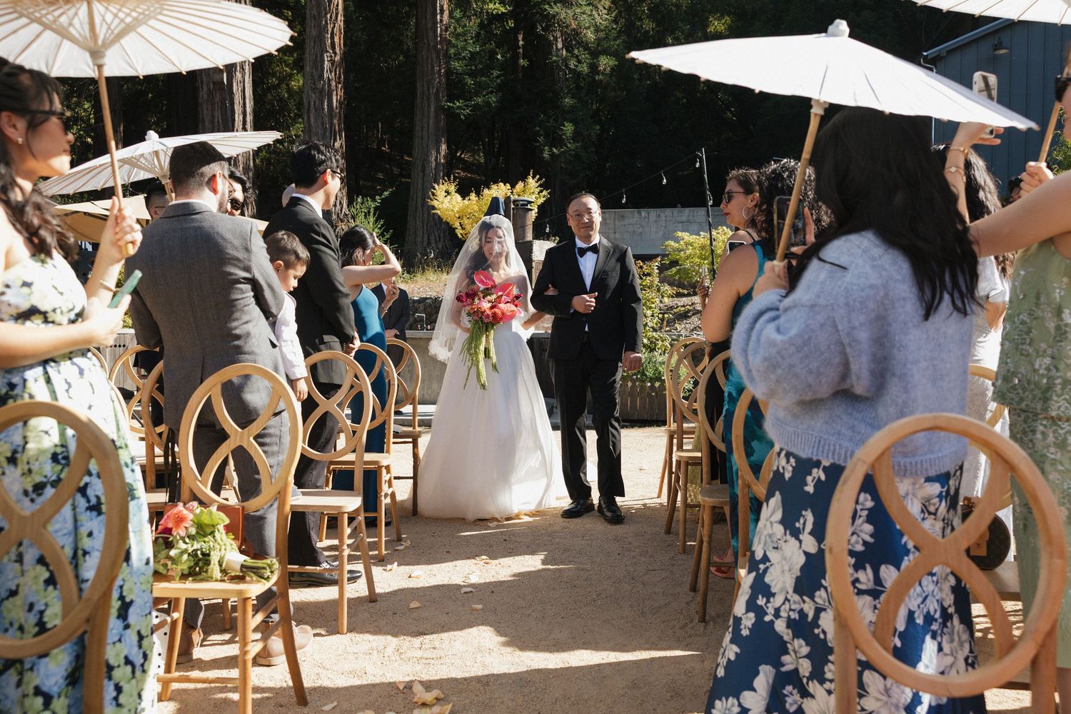 Outdoor garden party setting with white umbrellas and guests gathered around rustic wooden tables and chairs.
