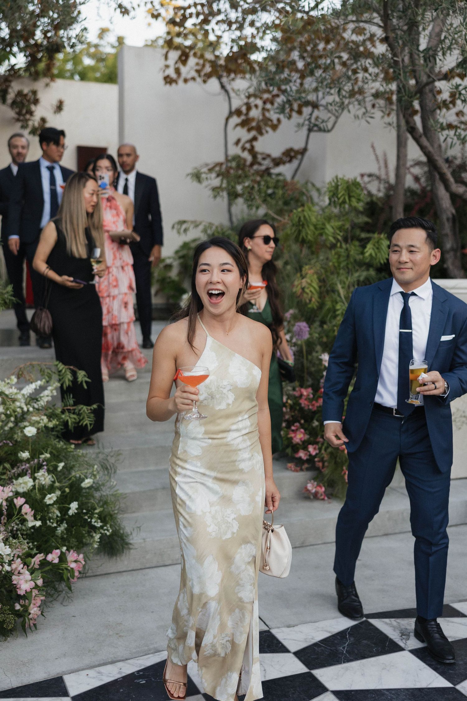 Wedding guests in formal attire walk along a flower-lined path at an outdoor reception.