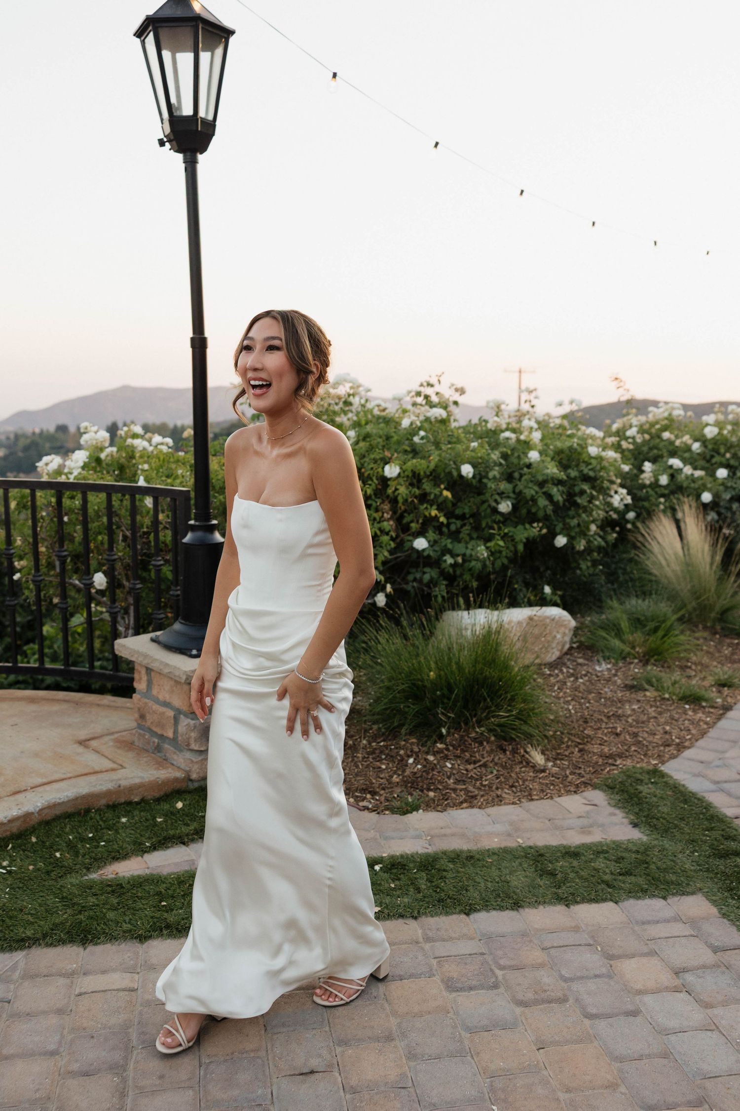 A bride in a simple white dress smiles while standing near a garden path with a vintage lamp post at dusk.