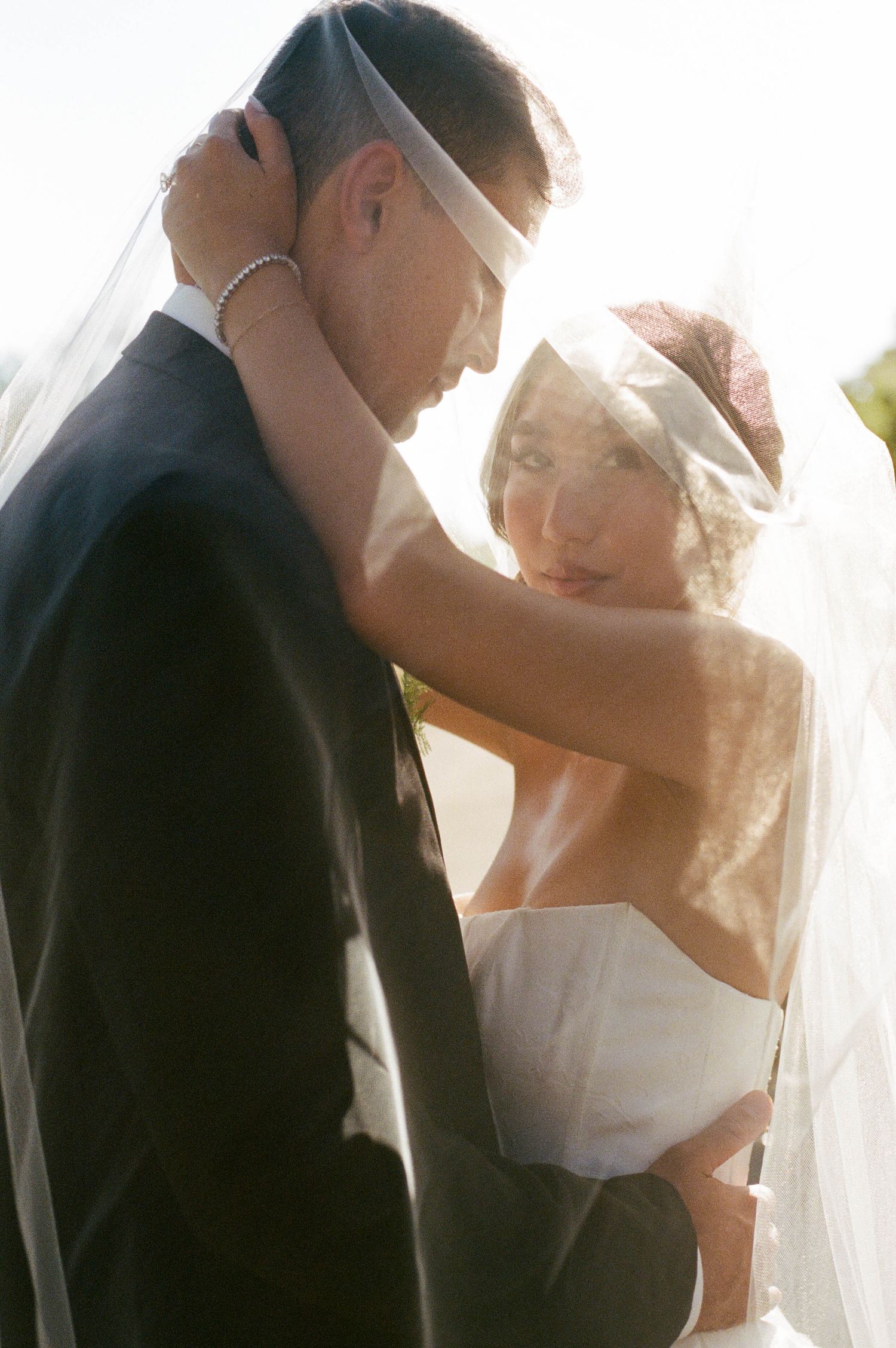 Intimate sunlit wedding moment captured through bride's veil as couple embraces.