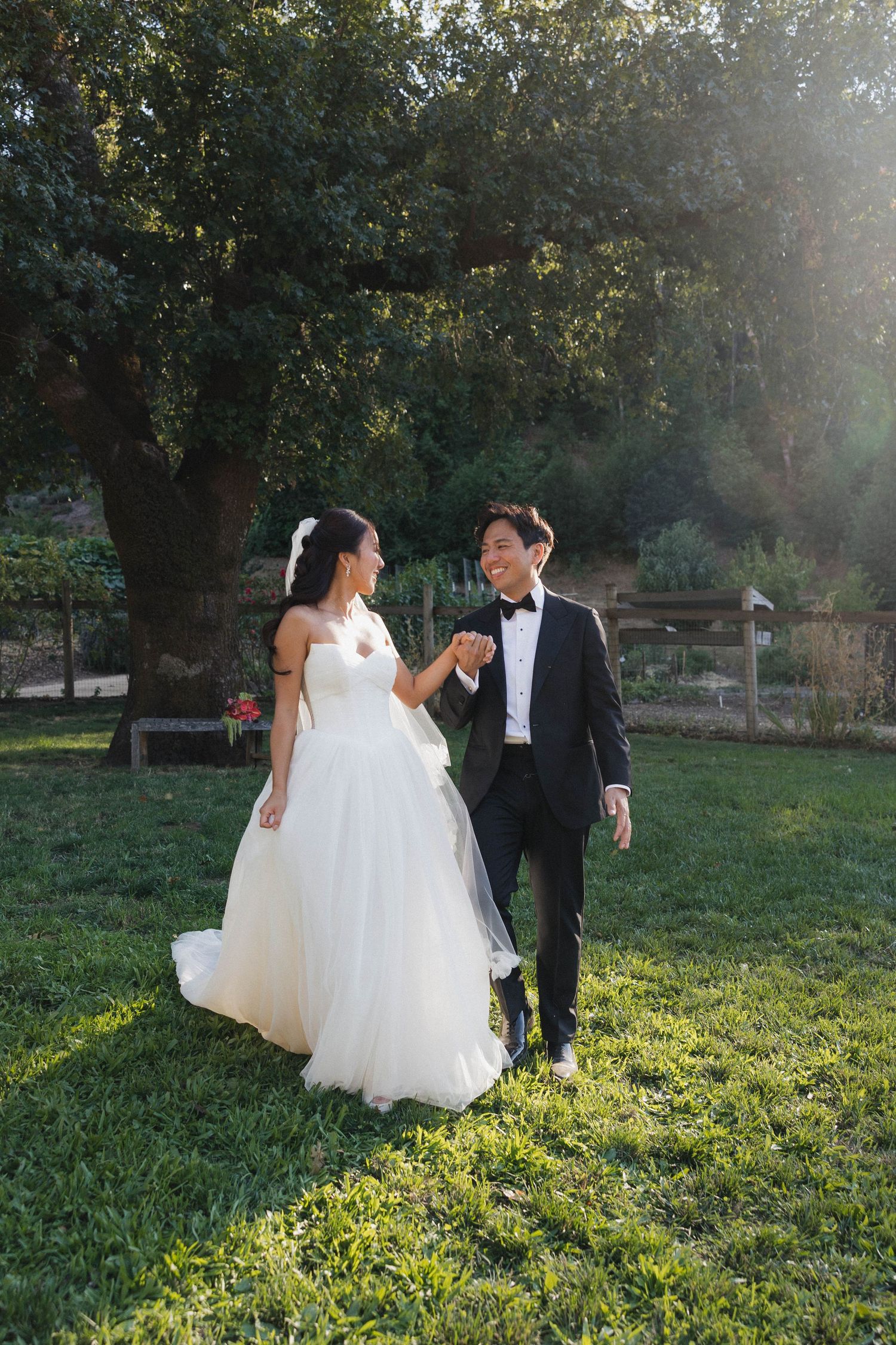 A couple in wedding attire walks hand in hand through a sunlit meadow at golden hour.