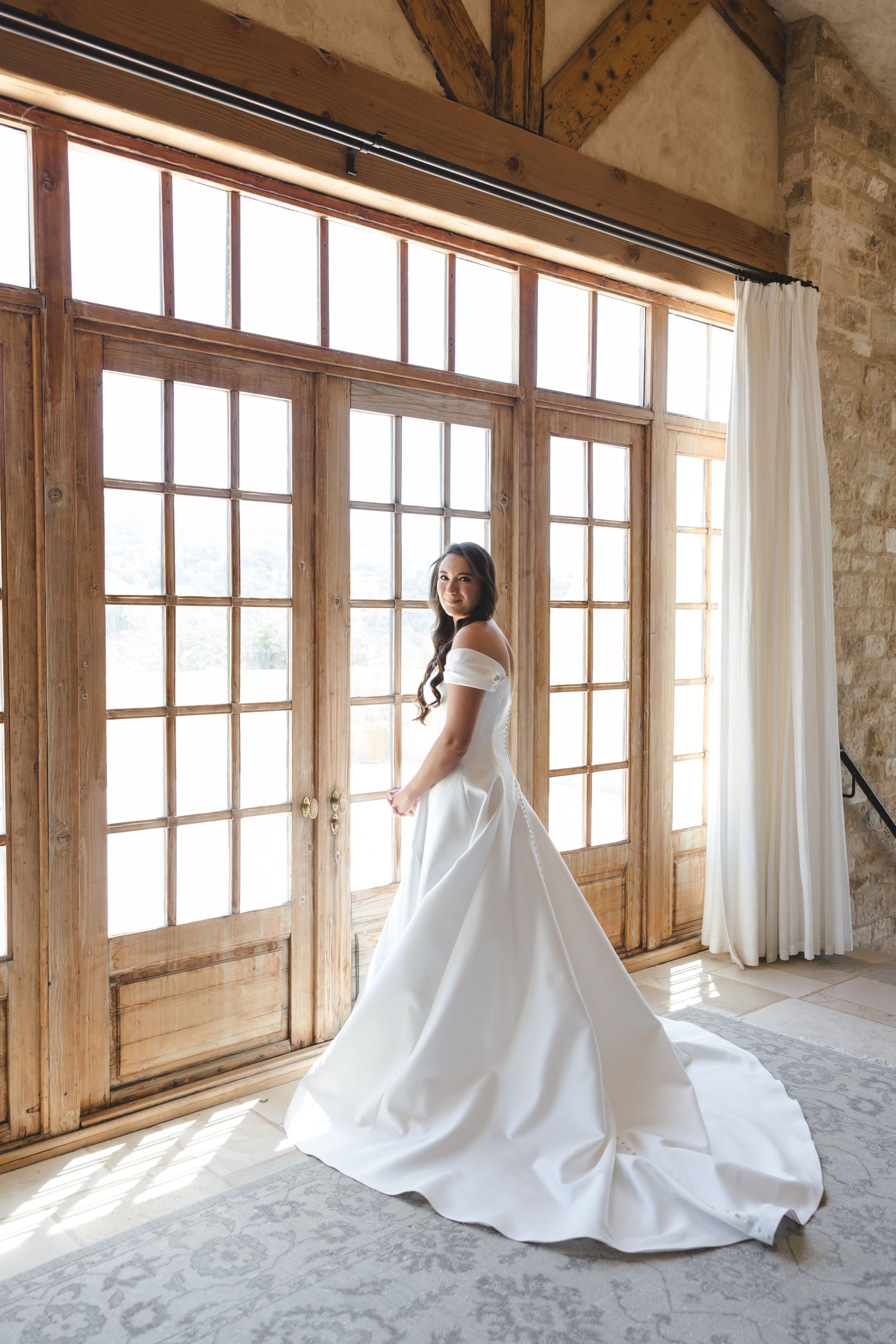 A stunning white wedding gown with long train is showcased in a sunlit room with rustic windows and exposed beams.