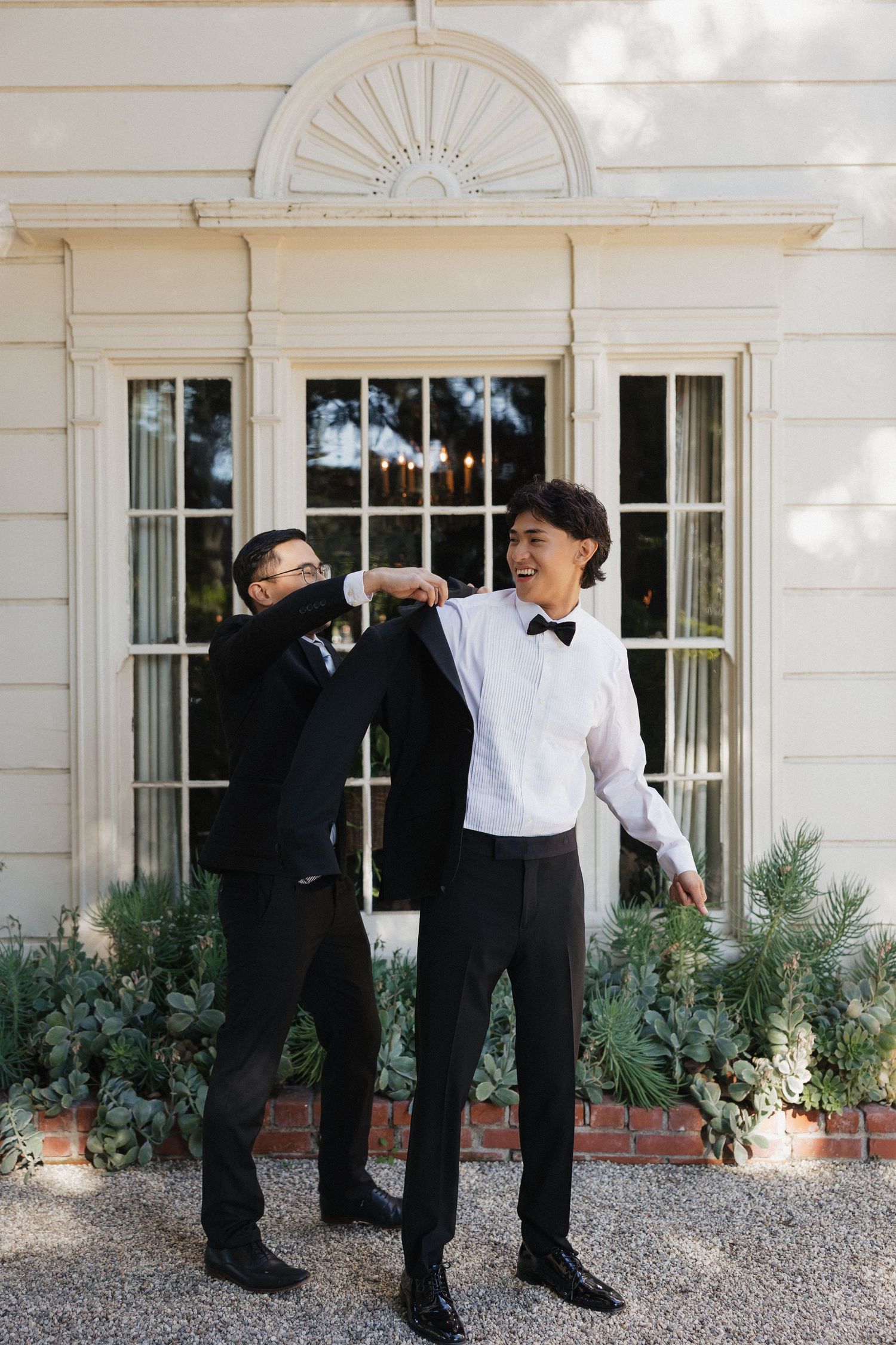 Groomsmen in black tuxedos pose playfully outside a white colonial style building.