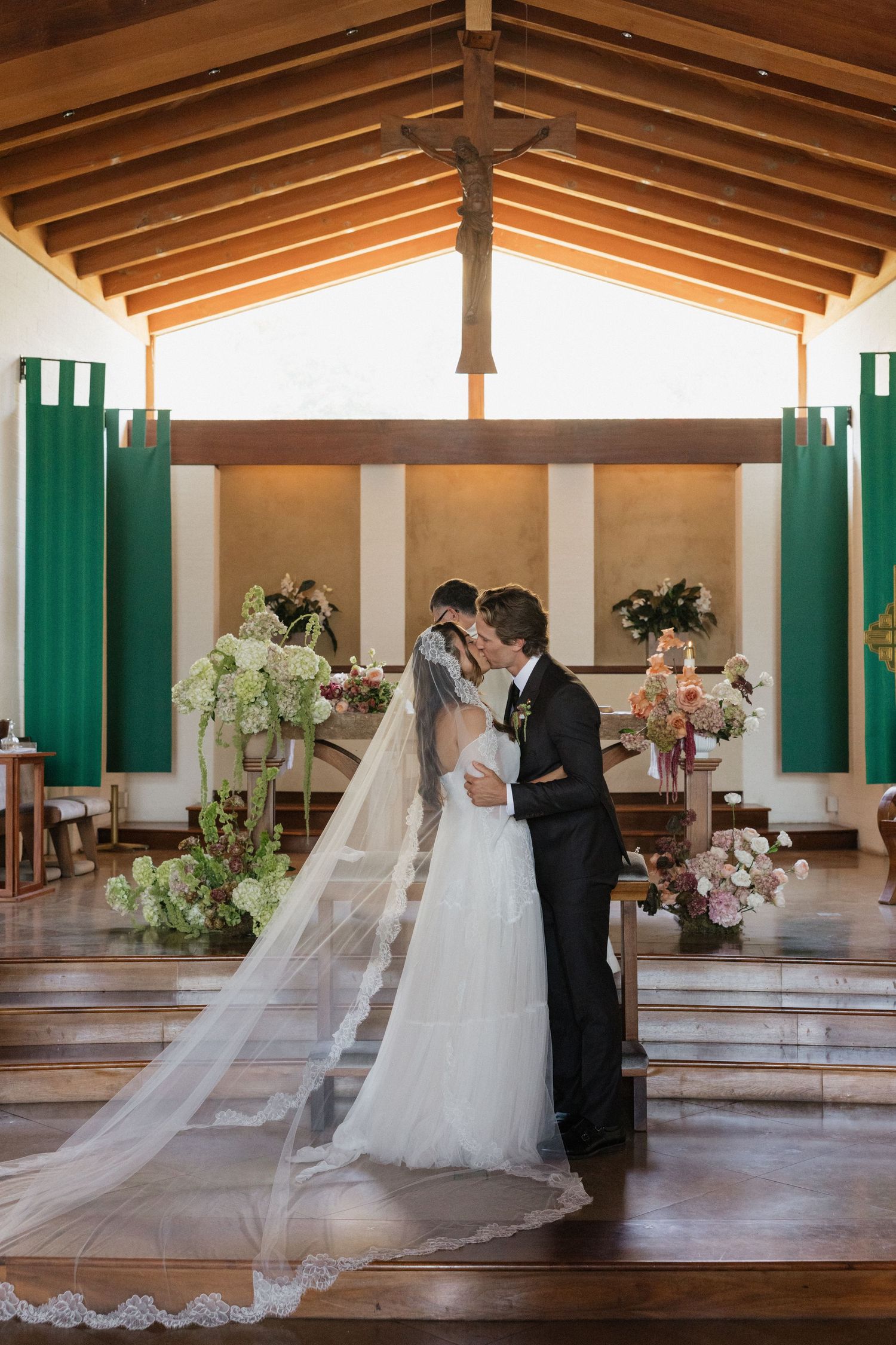 Bride and groom share first kiss at church altar adorned with floral arrangements and green banners.