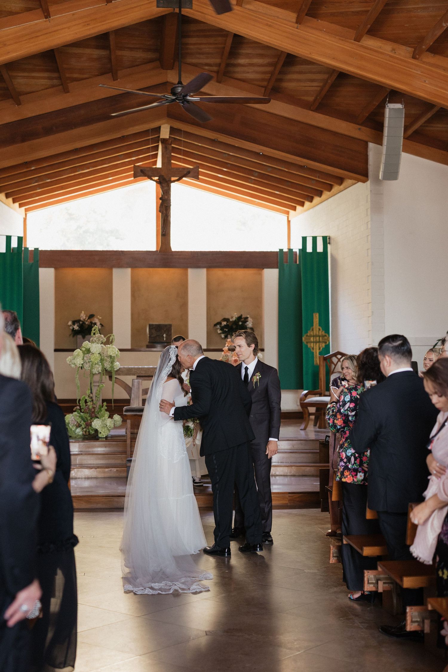 Bride in white gown and veil walks down church aisle with groom during wedding ceremony with wooden ceiling.