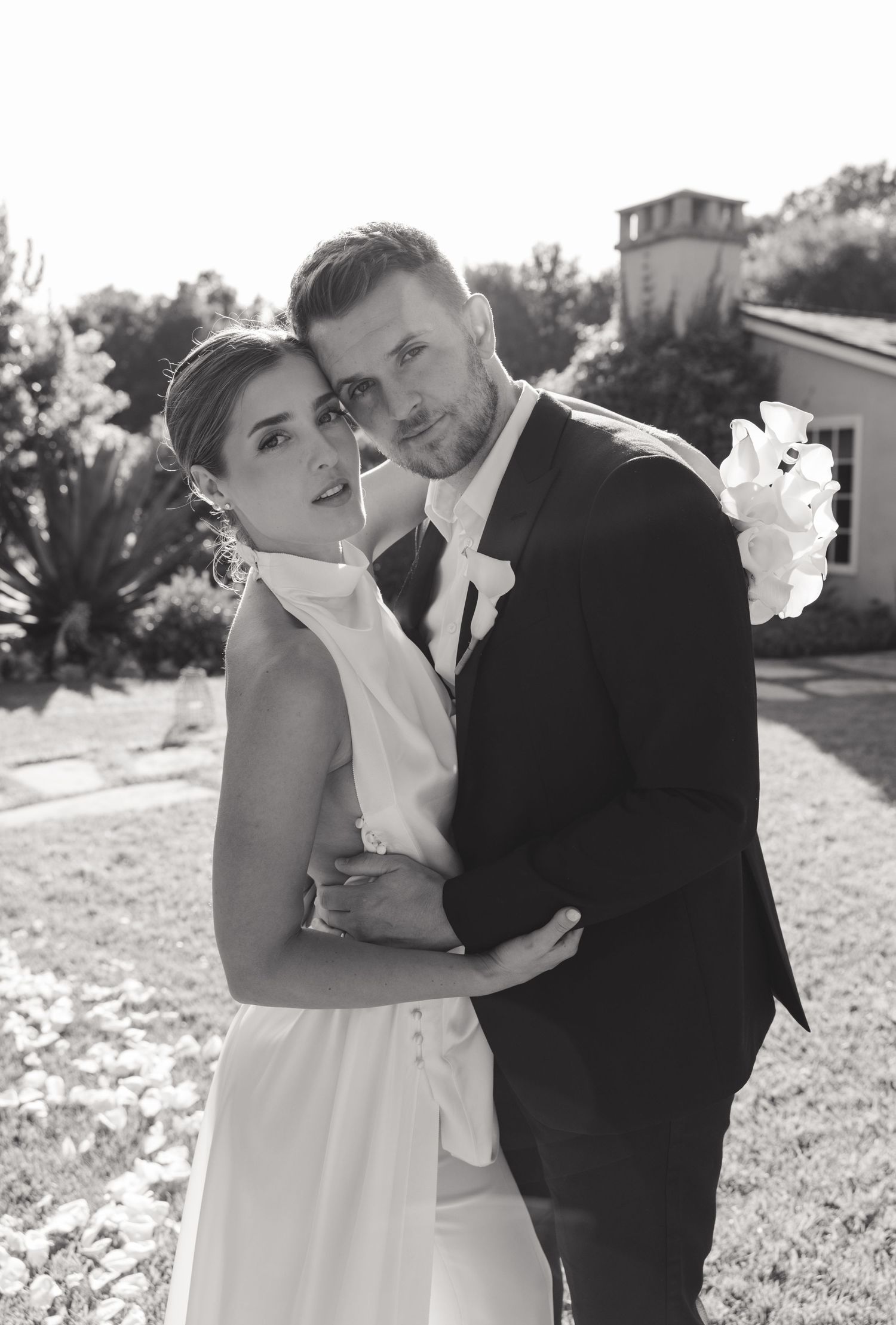 A bride in a high-neck white gown and groom in black suit embrace outdoors in a romantic black and white photograph.