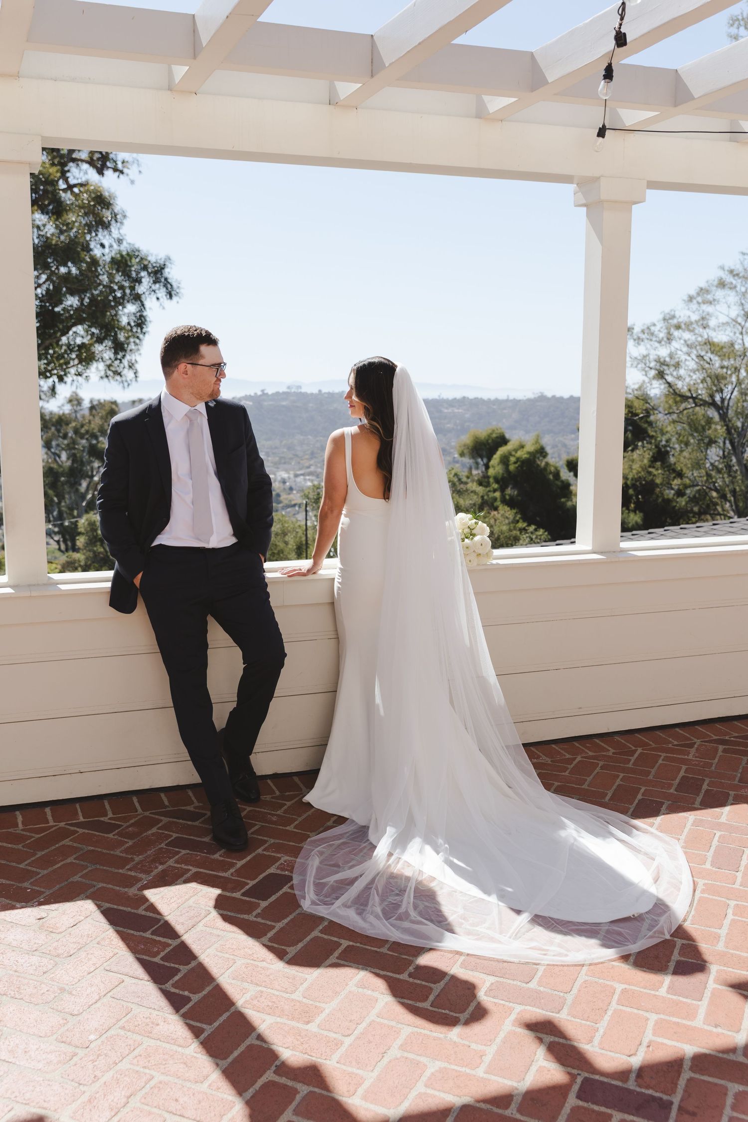 Newlyweds share an intimate moment on a bright brick veranda under a white pergola with mountain views in the distance.