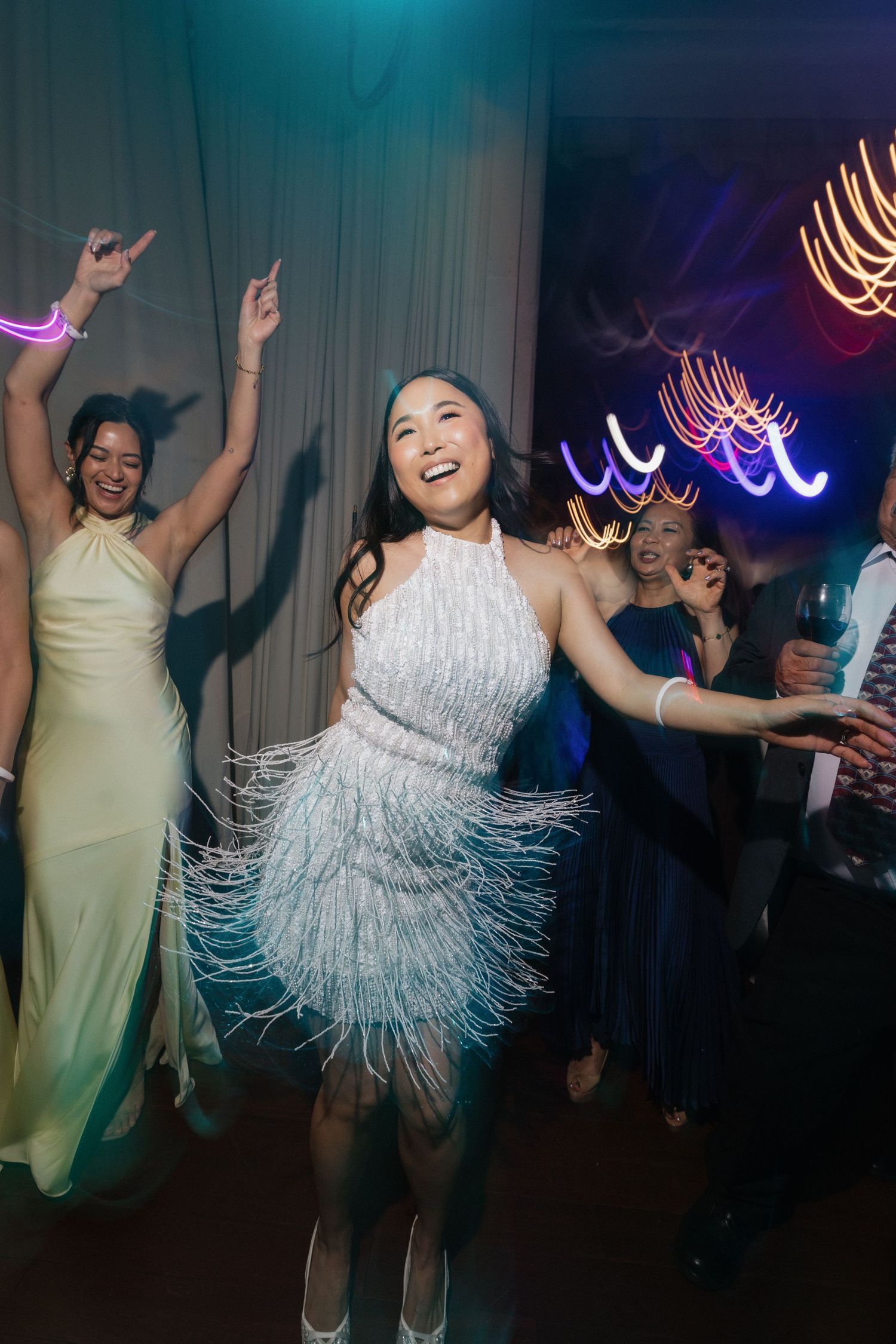 Woman in white fringe dress dancing joyfully at a party with colorful neon lights and guests celebrating.