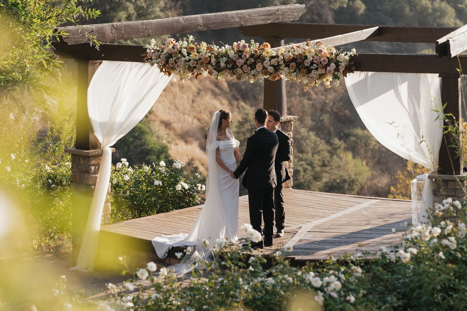 Elegant outdoor wedding ceremony setup with white fabric draping and floral arrangements along a mountain path.