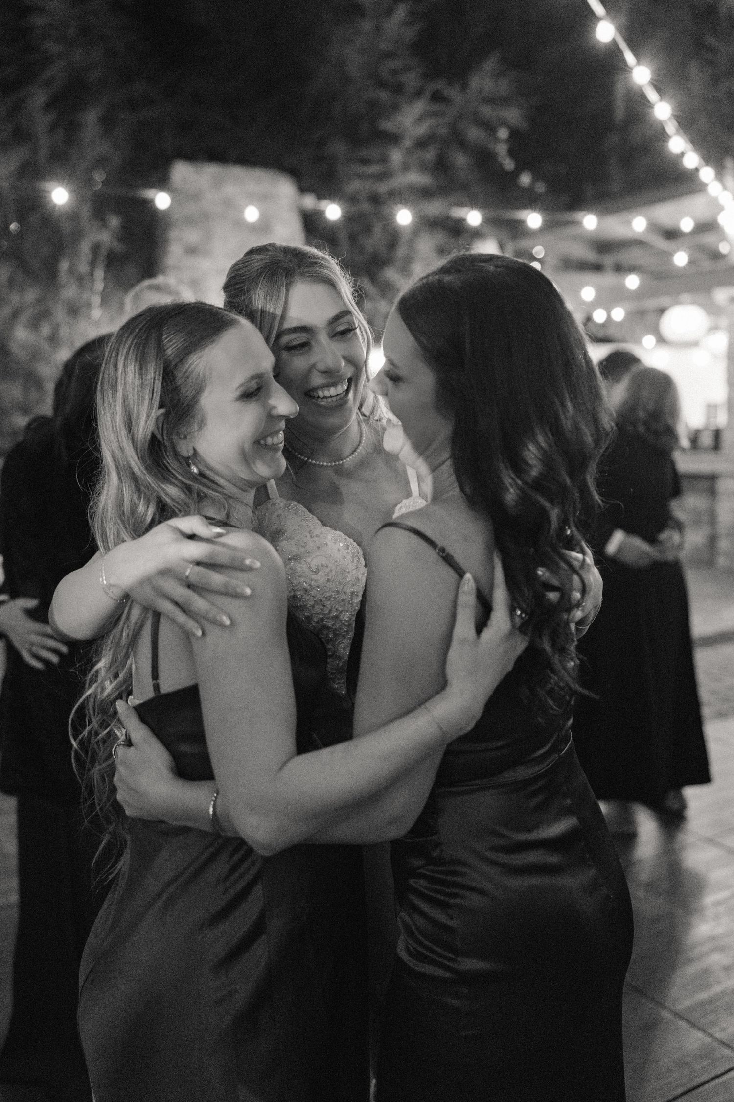 Three friends embrace and laugh together at an evening celebration under twinkling lights.
