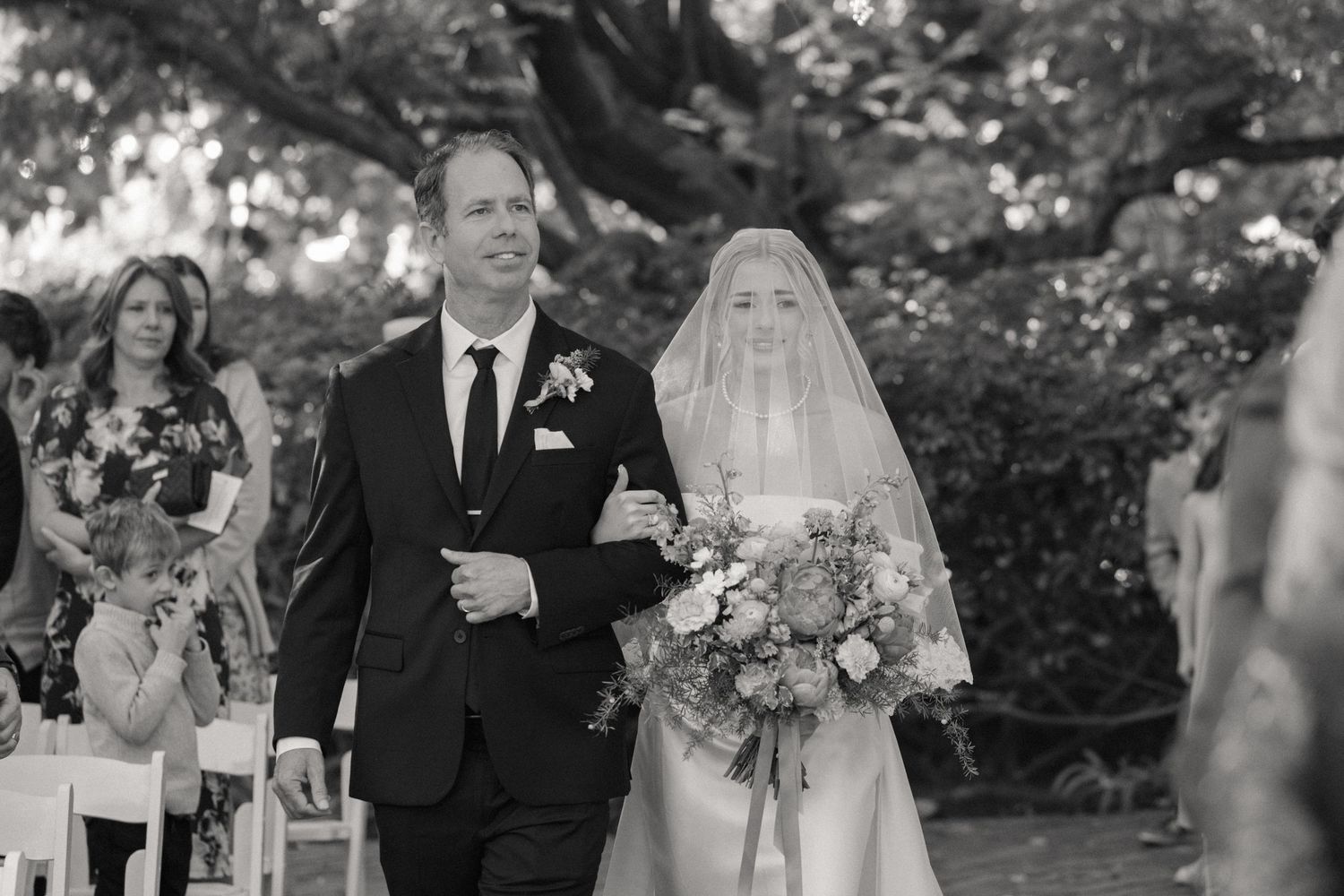 An outdoor wedding ceremony with floral arrangements and guests gathered under trees.